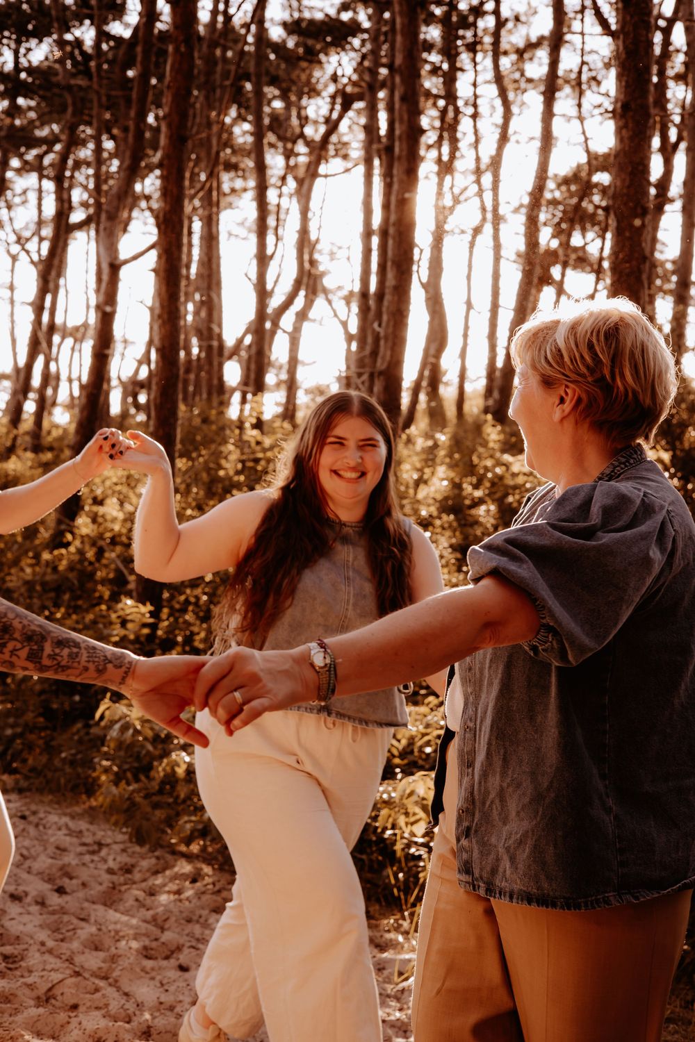Group of young women laughing and holding hands while dancing in a sun-drenched forest setting.