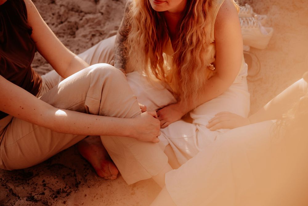 Two women sitting barefoot on sandy ground in warm golden sunlight near a forest edge.