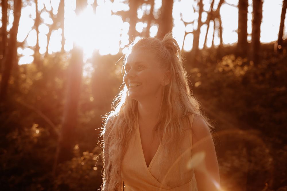 Smiling blonde woman with hair up standing in a radiant sunlit forest at golden hour.