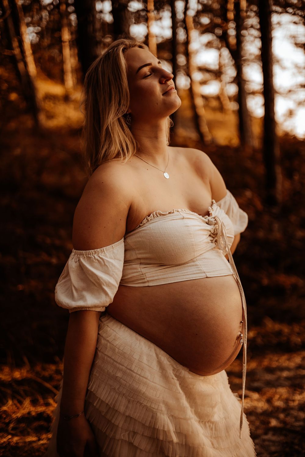 A pregnant woman in a white off-shoulder dress stands with eyes closed in a golden sunlit forest.