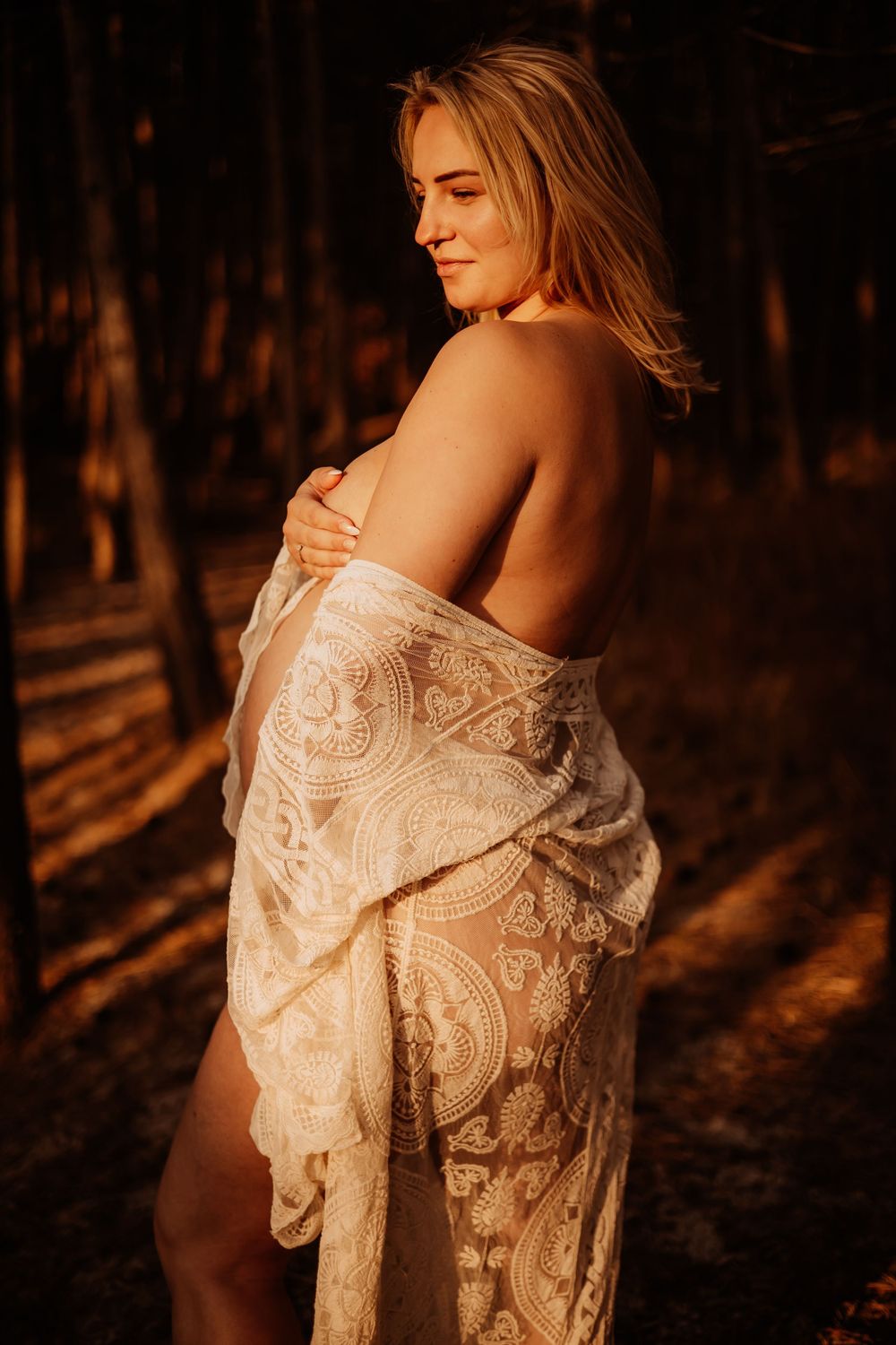 A pregnant woman draped in white lace fabric poses gracefully in a warm sunlit forest setting.
