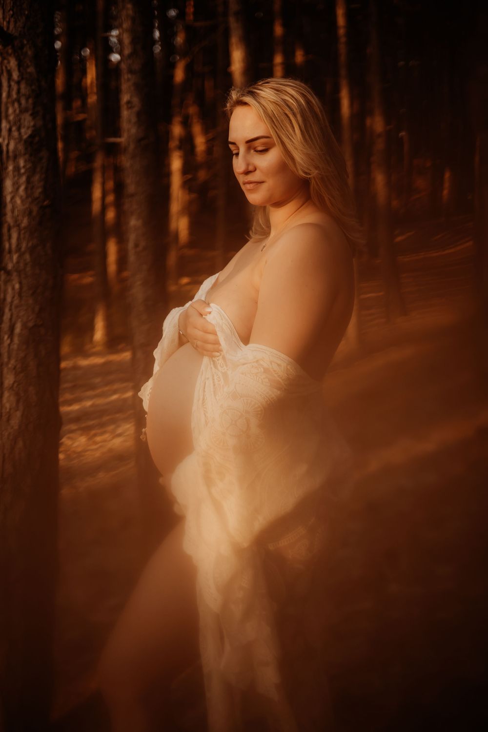 Pregnant woman in white lace robe cradling her baby bump in a sunlit pine forest at golden hour.