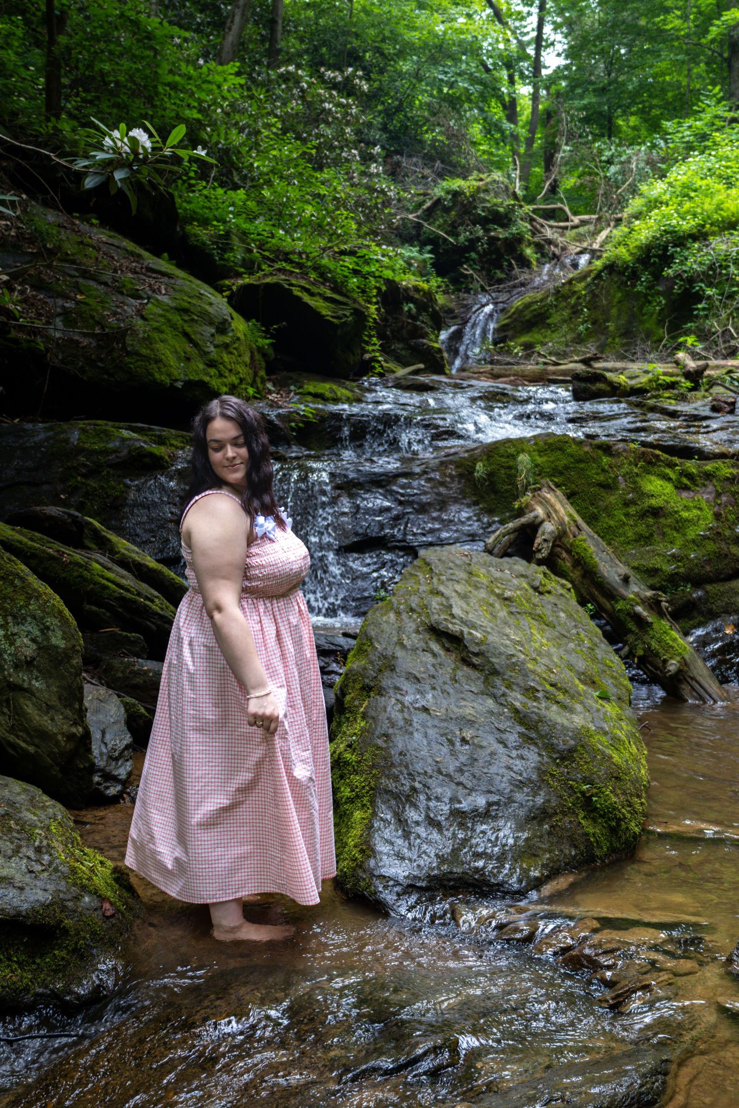 Woman in pink dress standing in a rocky stream beside moss-covered boulders with a waterfall in a lush forest.
