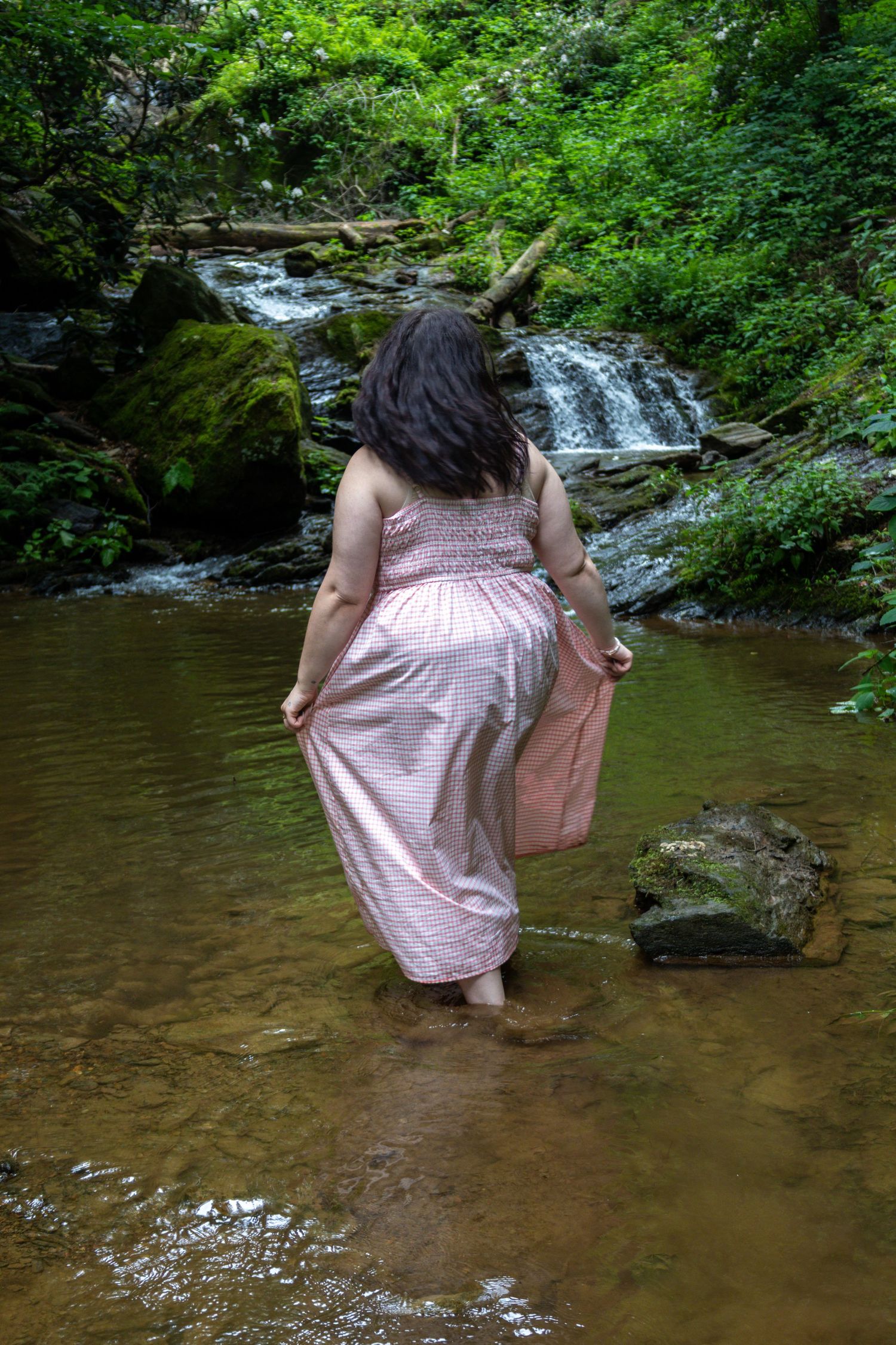 Woman in pink maxi dress wading in a serene forest stream with a lush green waterfall backdrop.