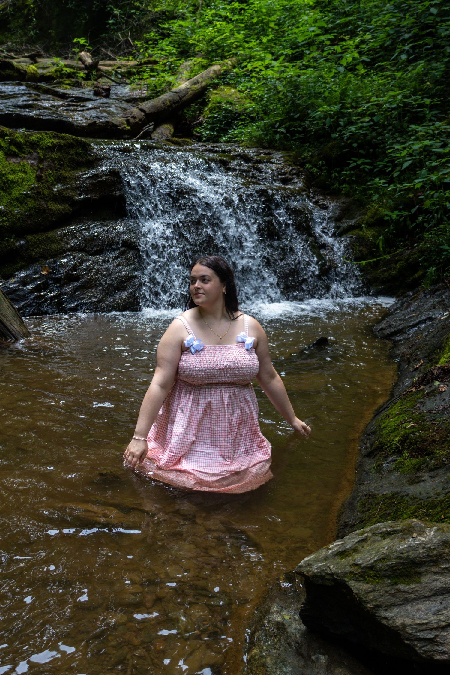 Woman in pink dress wading in a rocky stream with a lush green waterfall in the background.