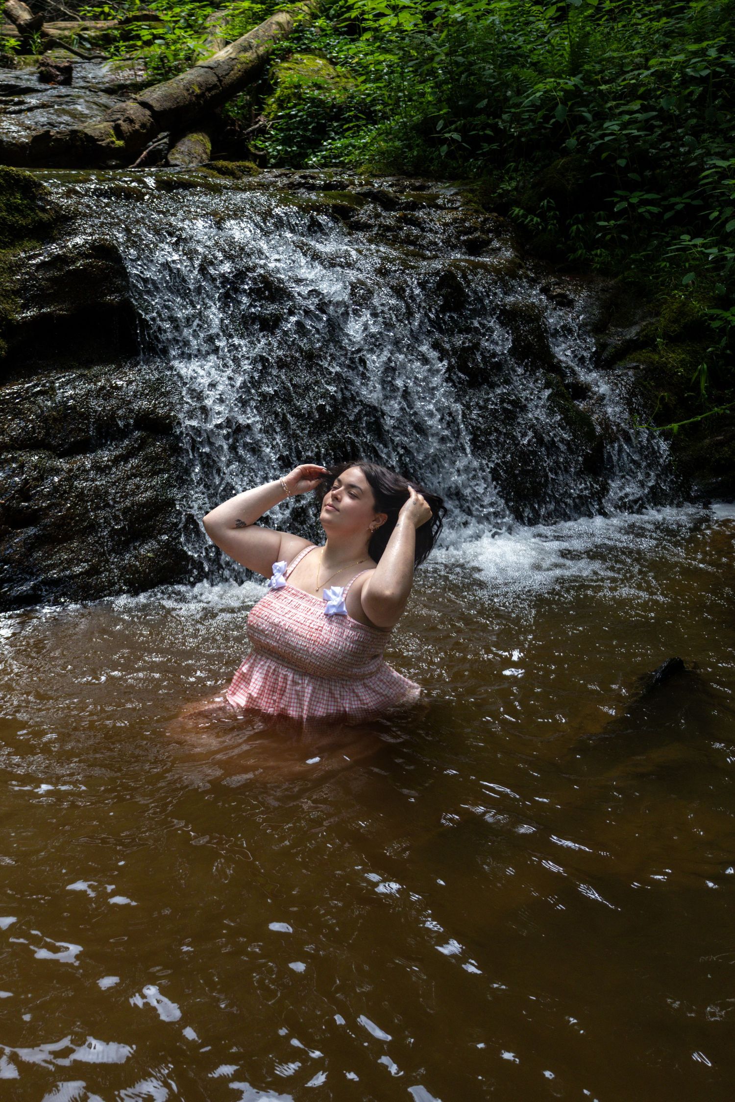 Woman in pink dress wading in a rocky forest waterfall pool, surrounded by lush green vegetation.