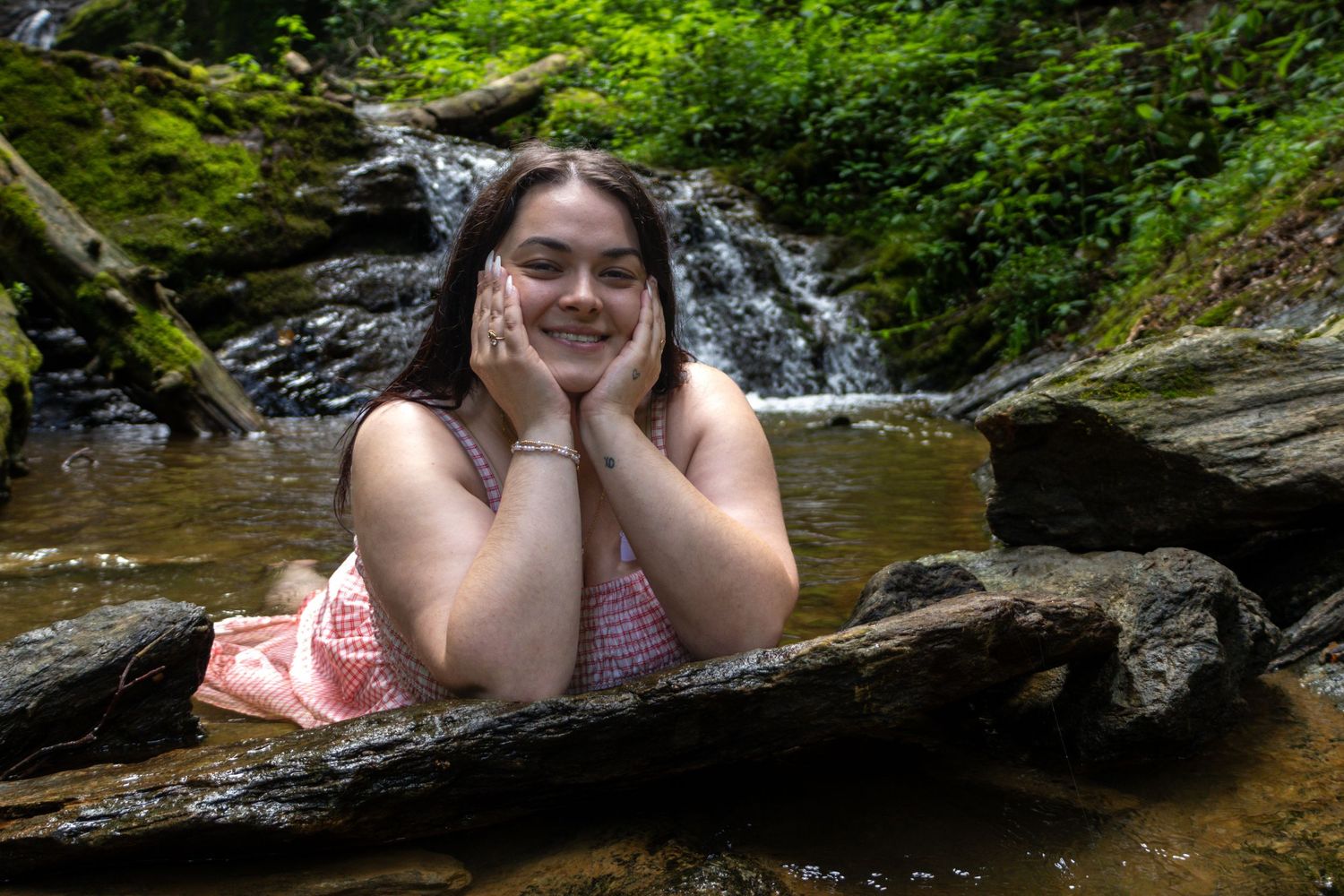 Young woman smiling by a woodland waterfall, resting on rocks in a serene forest stream setting.