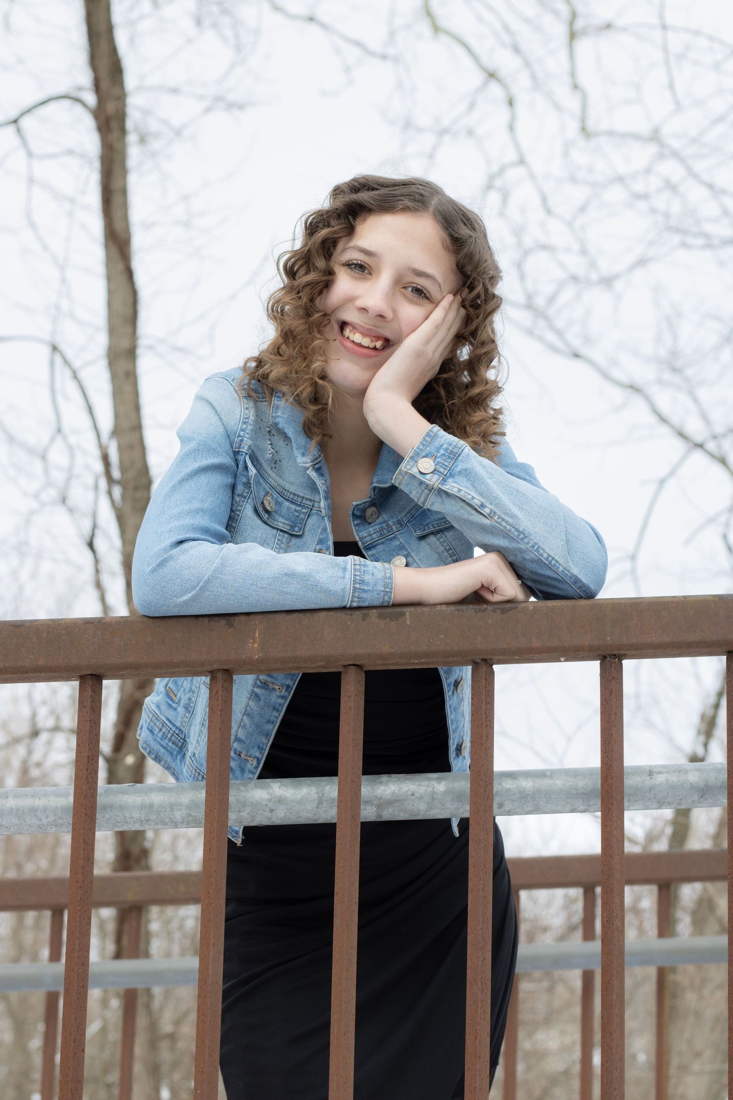 Smiling young woman with curly hair in denim jacket leaning on wooden railing outdoors.
