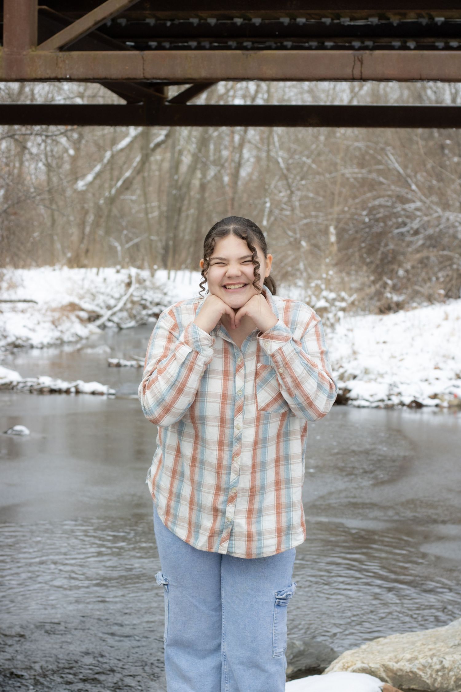Young woman in plaid shirt and jeans smiling by a snowy creek under a bridge in winter.