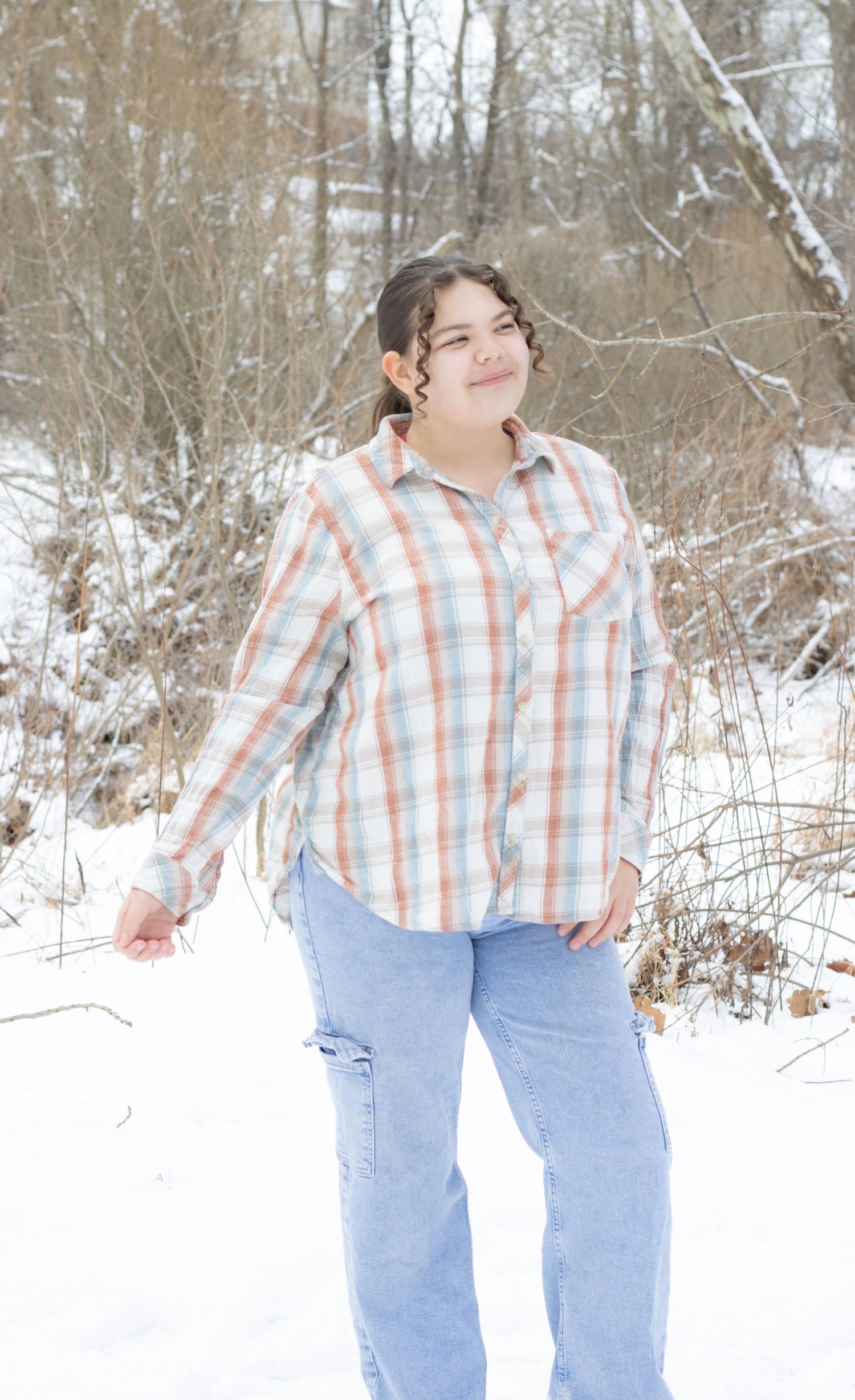 Young woman in plaid shirt and jeans smiling outdoors in snowy winter landscape with bare trees.