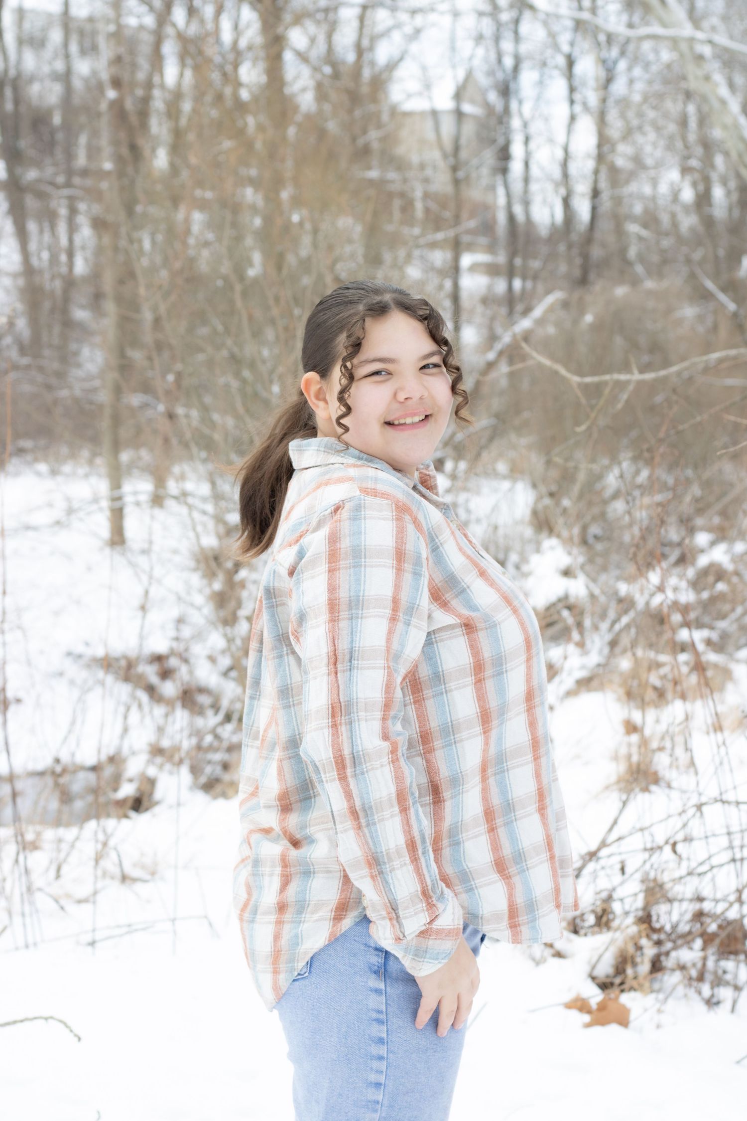 Young girl in plaid shirt and jeans smiling outdoors in a snowy winter landscape with bare trees.