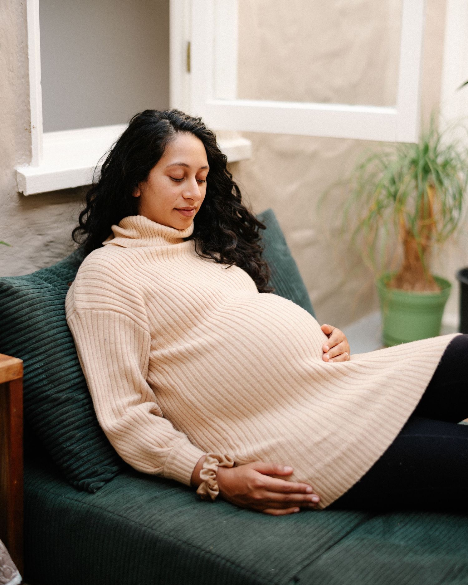 Pregnant woman in beige turtleneck sweater dress relaxing on green sofa, cradling baby bump with eyes closed.