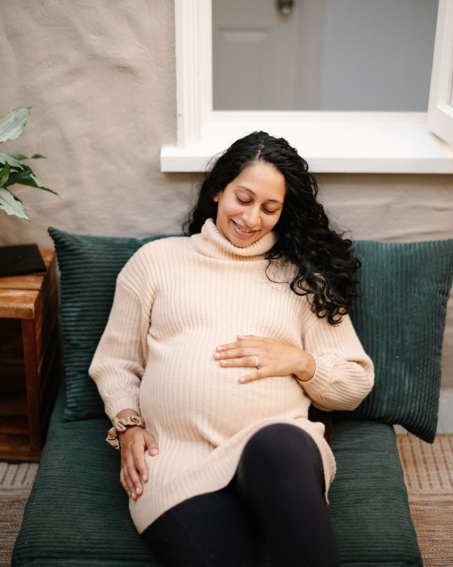 Smiling pregnant woman in beige turtleneck sweater cradling baby bump while relaxing on green couch.