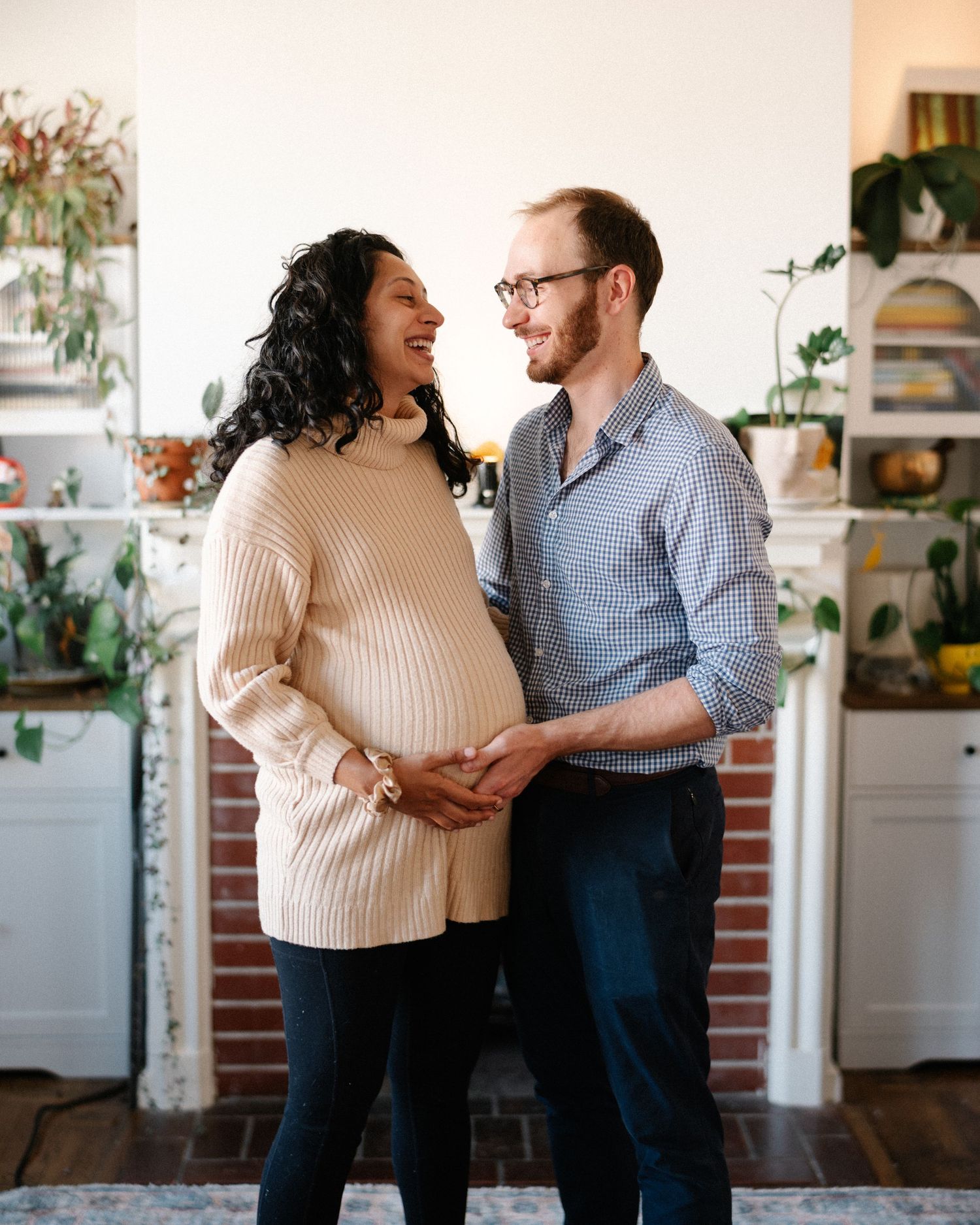 Happy interracial couple sharing a joyful moment, hands on pregnant belly, standing in a plant-filled home.