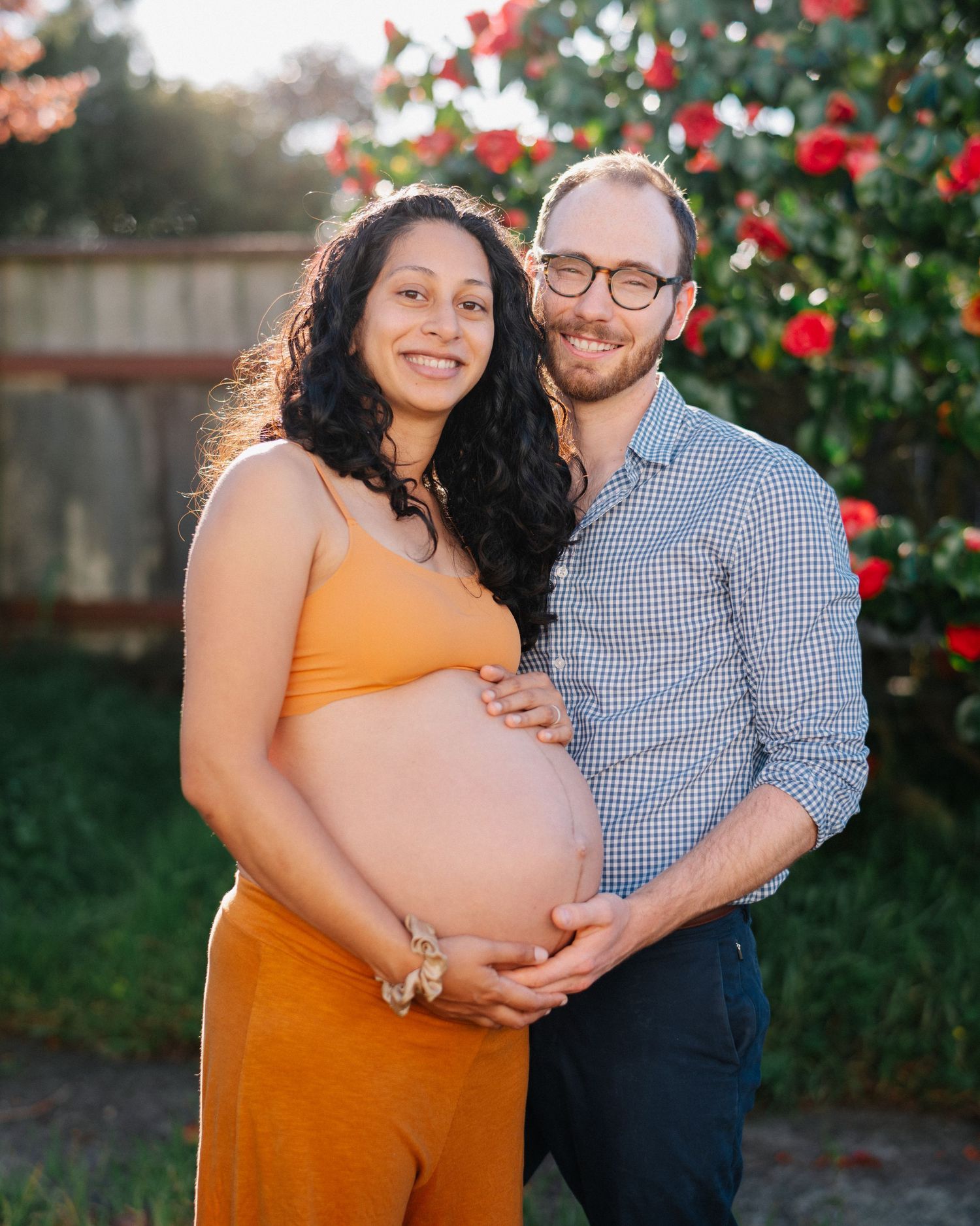 Couple posing outdoors for maternity photo with red roses backdrop, man embracing pregnant woman in orange dress.
