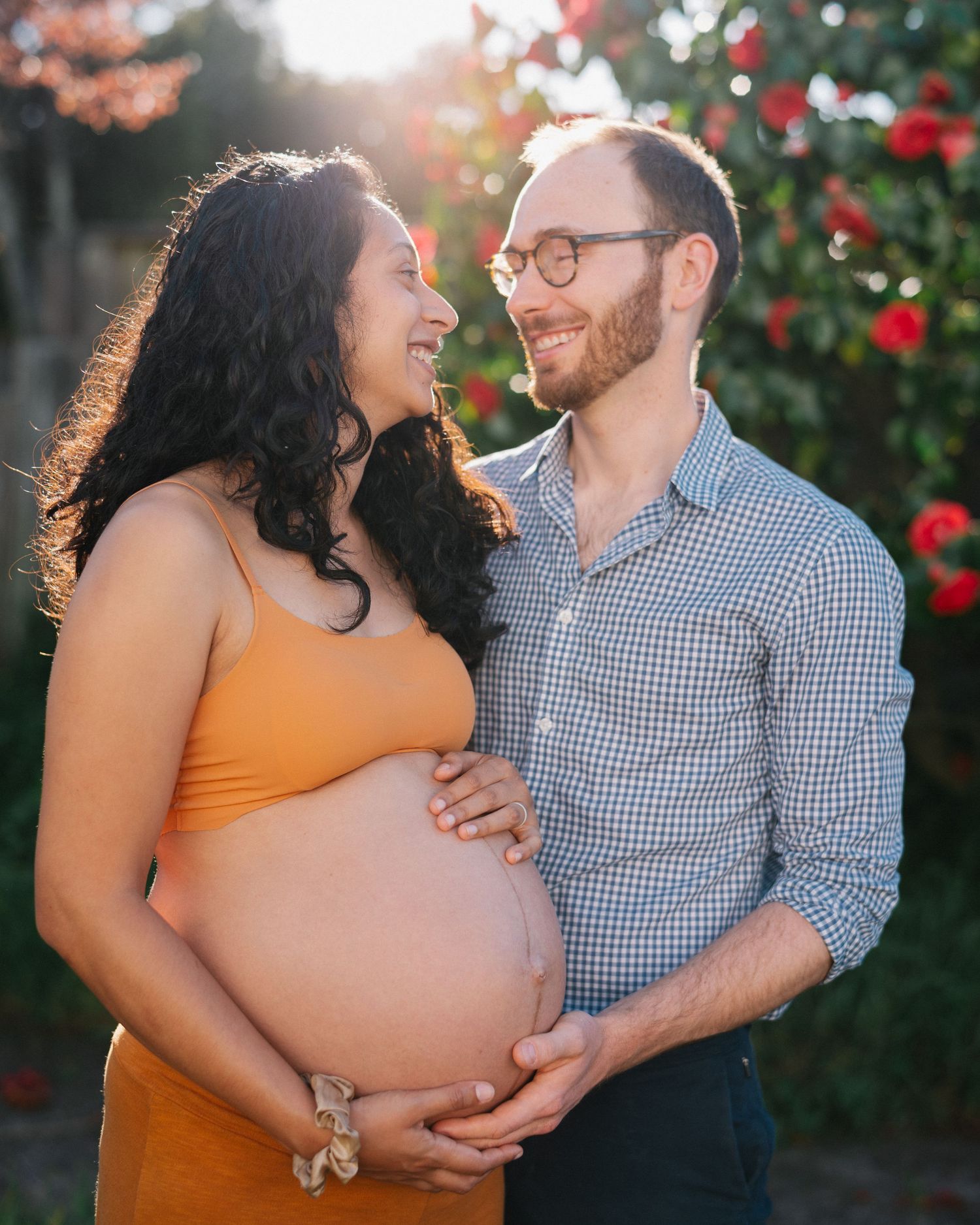 Glowing pregnant couple holding baby bump outdoors at golden hour with red roses blooming behind them.