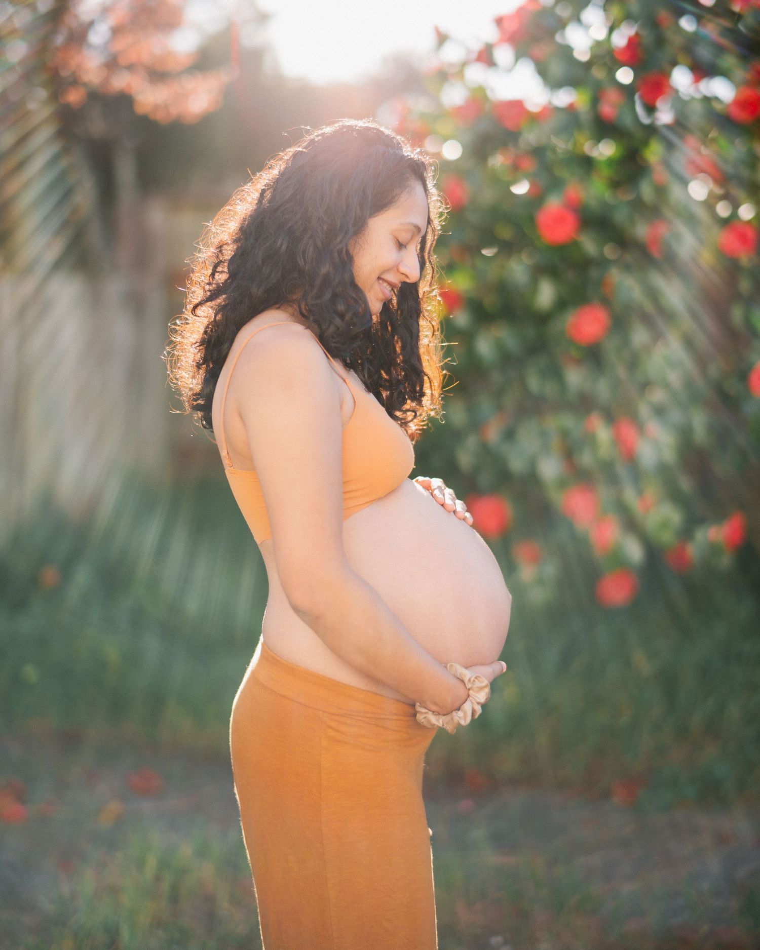 Pregnant woman in orange crop top and pants holding baby bump outdoors with red flowers in golden sunlight.