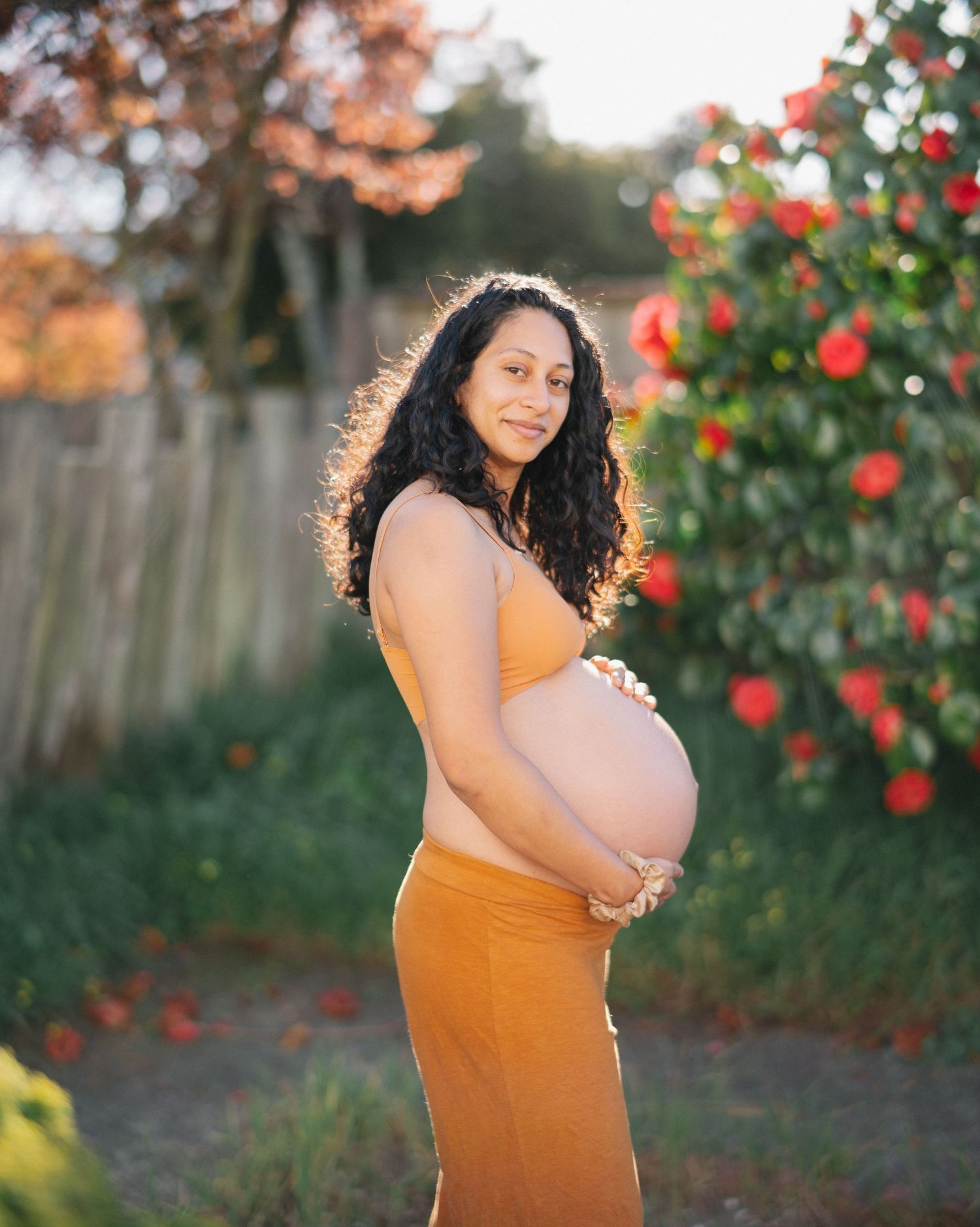 Pregnant woman in orange dress cradling baby bump outdoors with blooming red roses in golden hour light.