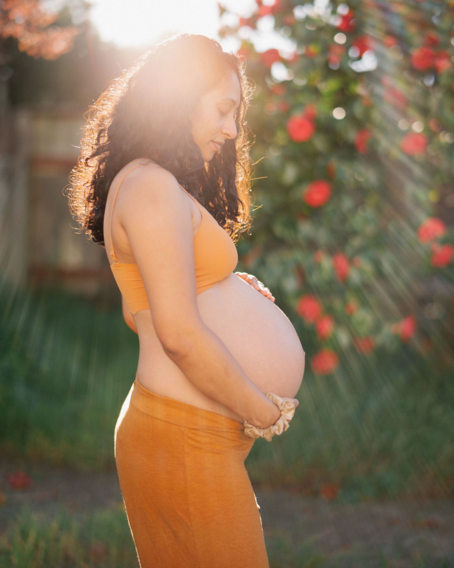 Side profile of pregnant woman in mustard outfit cradling belly outdoors in warm golden hour sunlight.