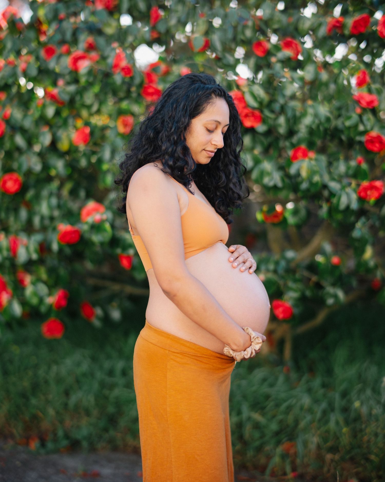 Pregnant woman in orange outfit cradling her baby bump in front of blooming red roses outdoors.