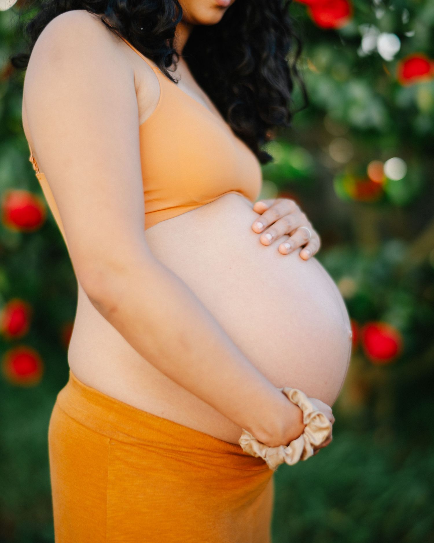 Pregnant woman in orange crop top and skirt cradling baby bump outdoors with red flowers in background.