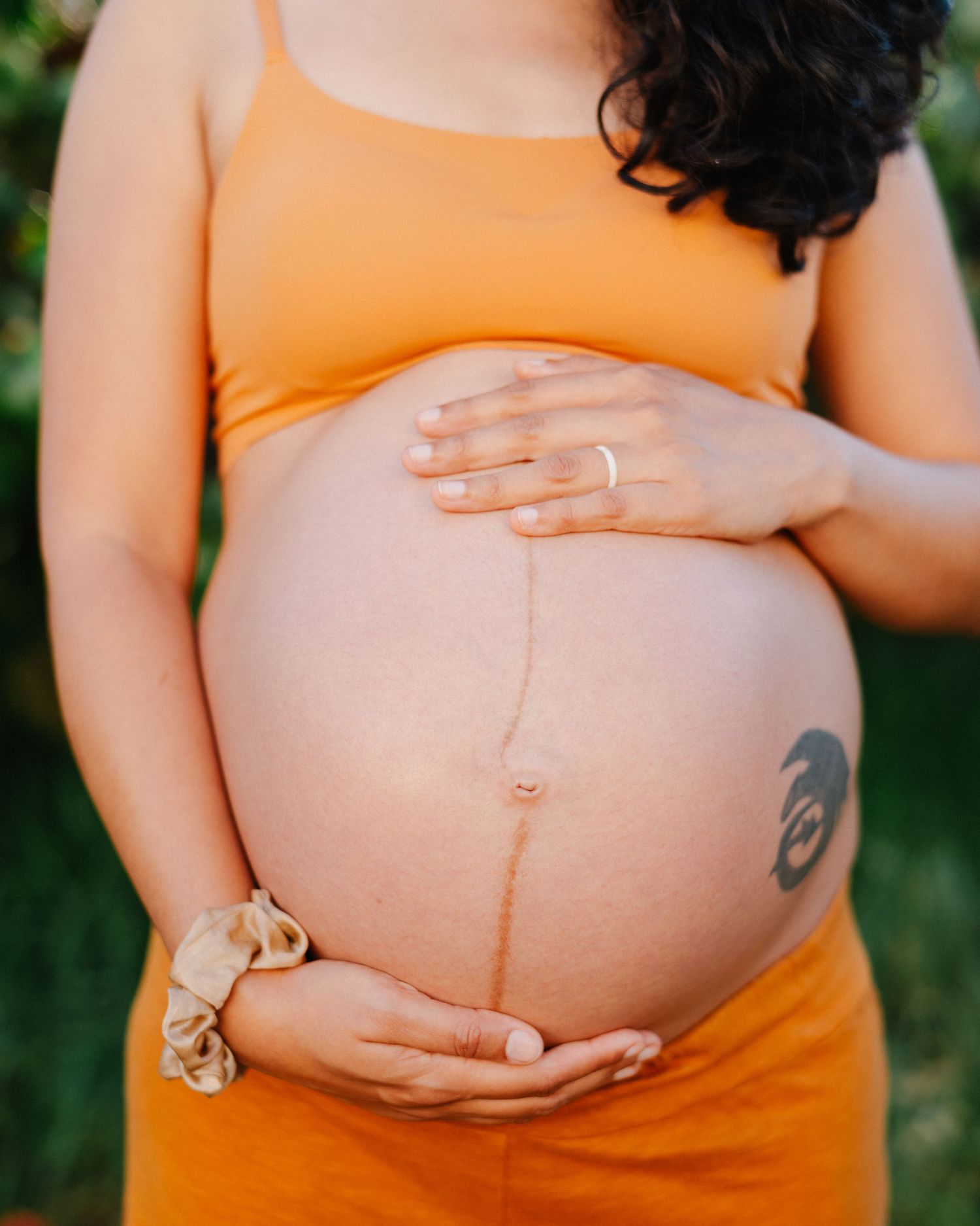 Pregnant woman in orange dress cradling her baby bump outdoors, showing linea nigra and a tattoo.
