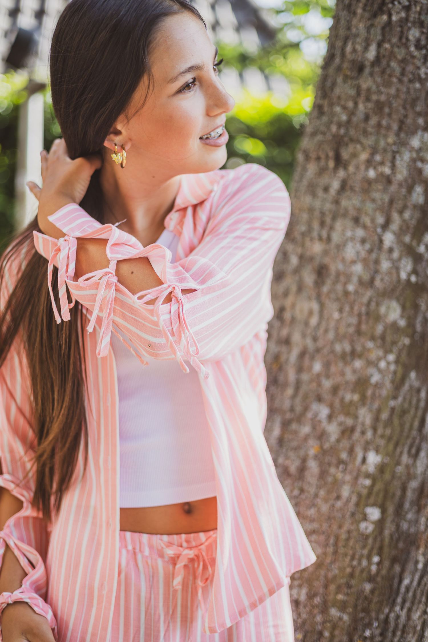Young woman in pink striped matching set with bow details, posing outdoors near a tree in natural light.
