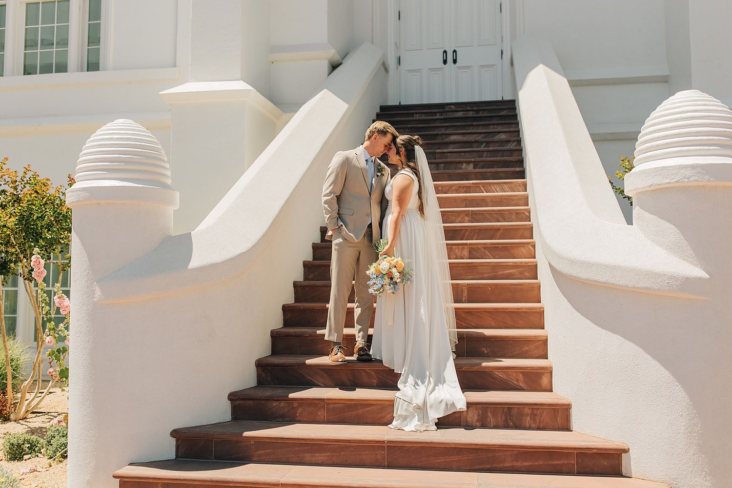 Bride and groom kissing on white stucco staircase outside elegant wedding venue on sunny day.