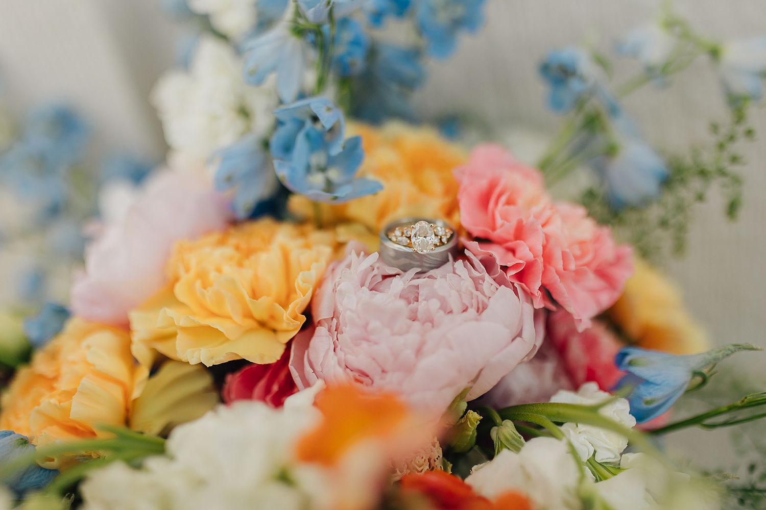 Wedding rings resting on a colorful bridal bouquet featuring pink peonies, yellow, orange, and blue flowers.