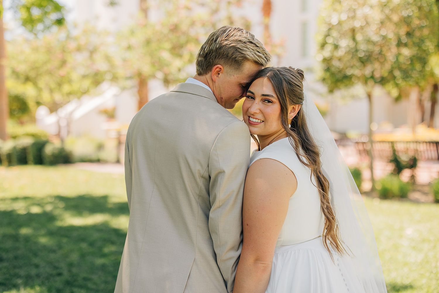 Bride smiling over shoulder while groom nuzzles her hair outdoors in a lush green garden setting.