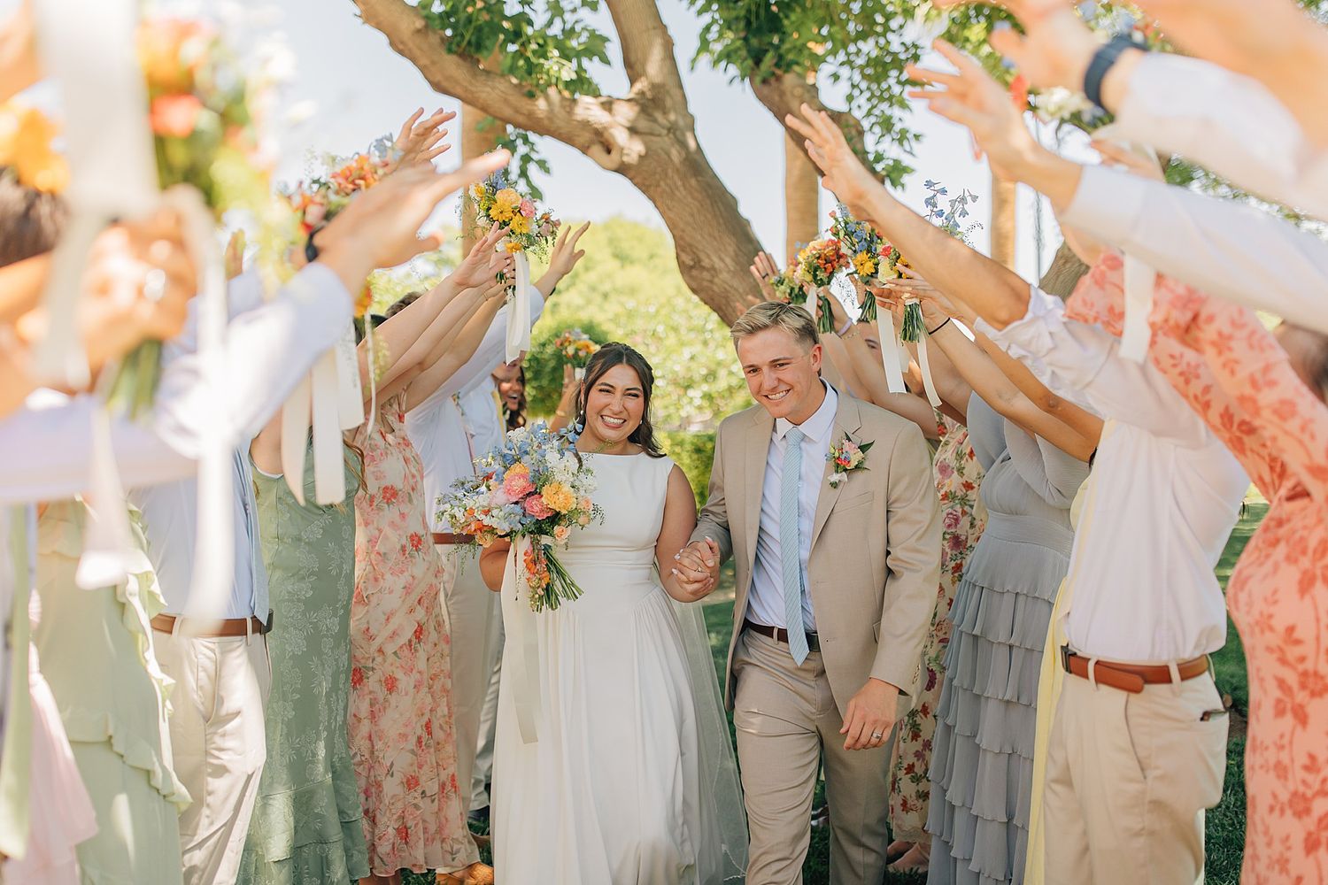 Bride and groom walk through floral arch tunnel surrounded by cheering wedding guests outdoors.