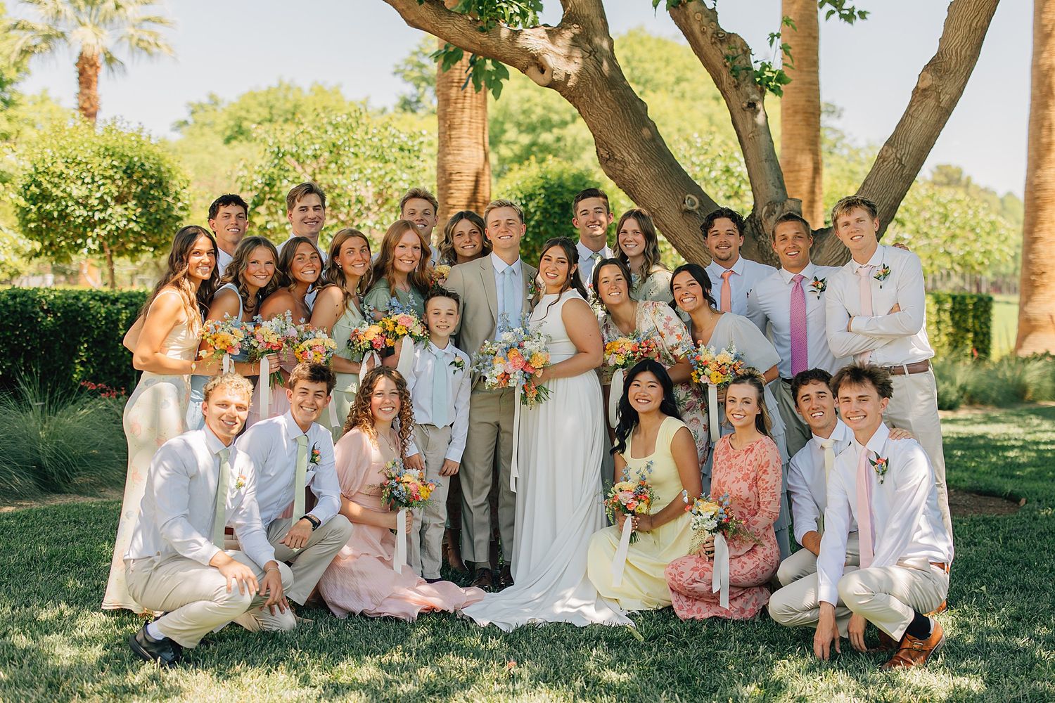 Large wedding party poses outdoors under trees, bride and groom centered among guests in pink and white attire.