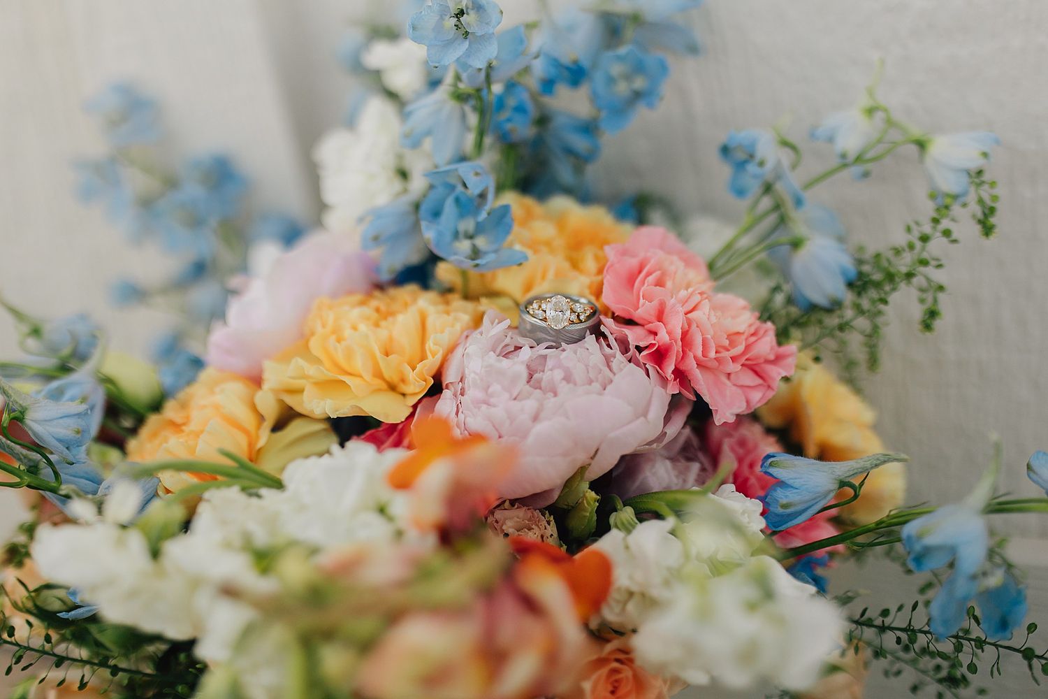 Colorful wedding bouquet with pink peonies, yellow and orange roses, blue delphinium, and a diamond engagement ring resting on top.