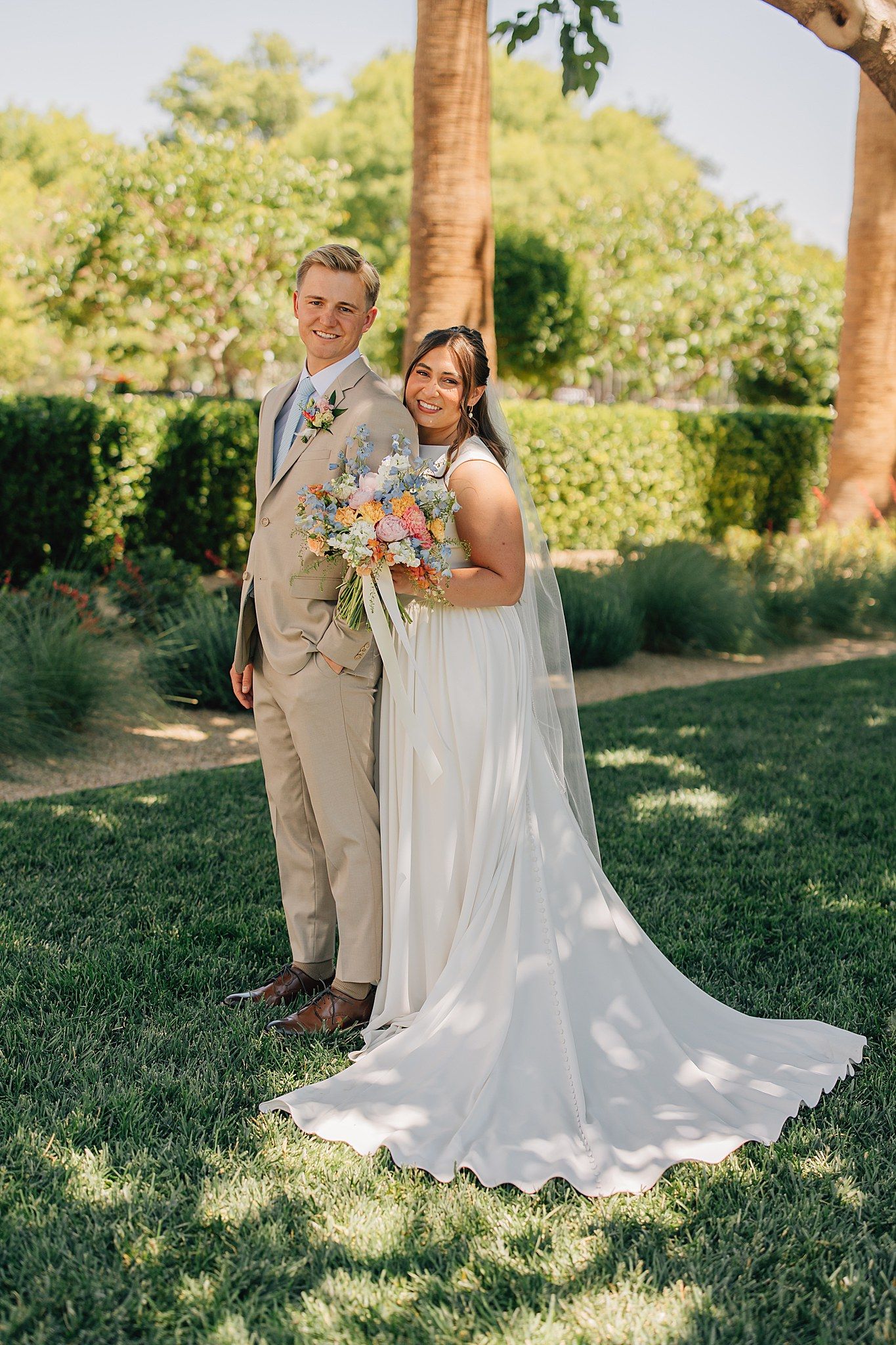 A bride in a white gown holding a colorful bouquet stands with a groom in a tan suit on a green lawn.