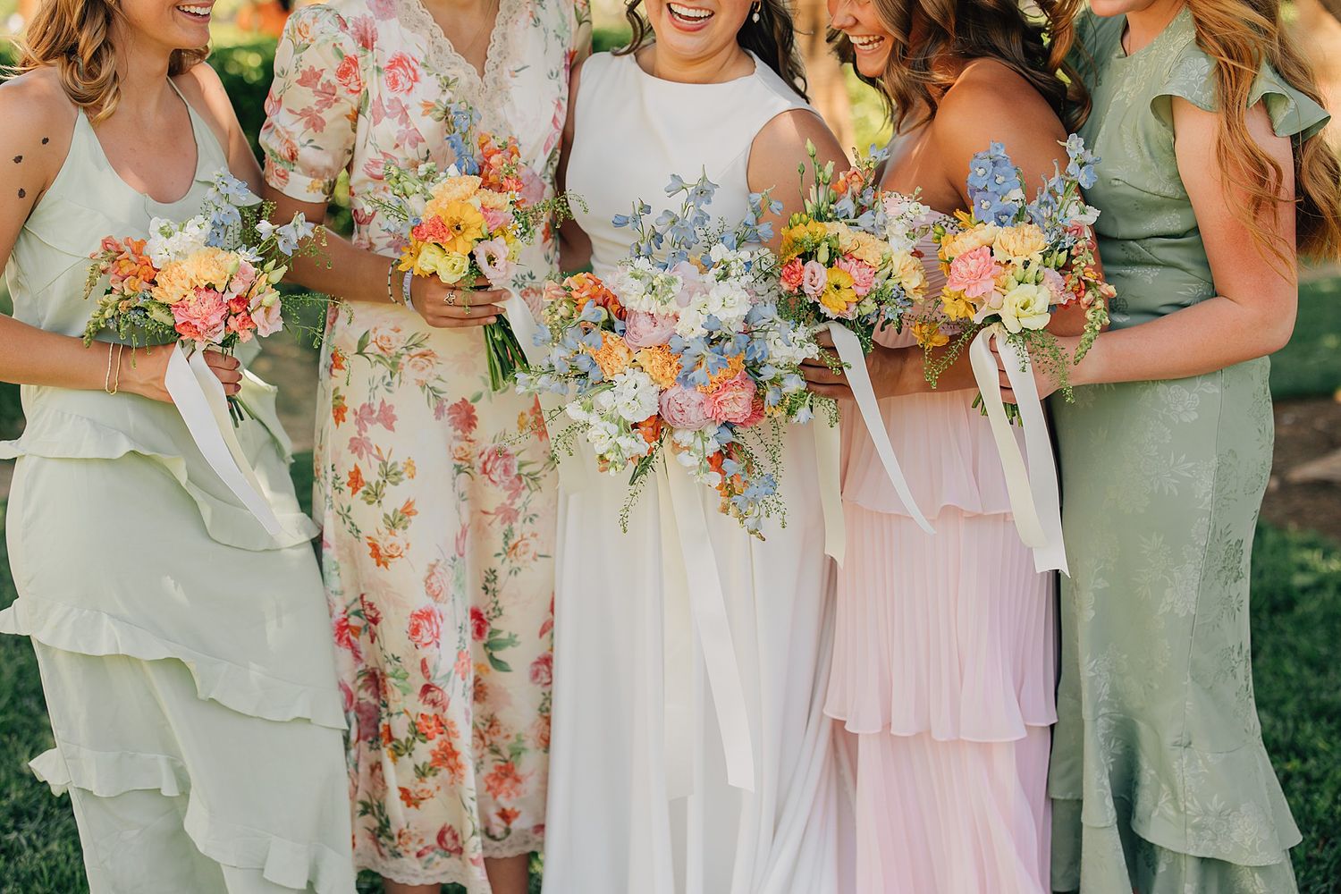 Bride and bridesmaids in mismatched sage green and floral dresses holding colorful wildflower bouquets outdoors.