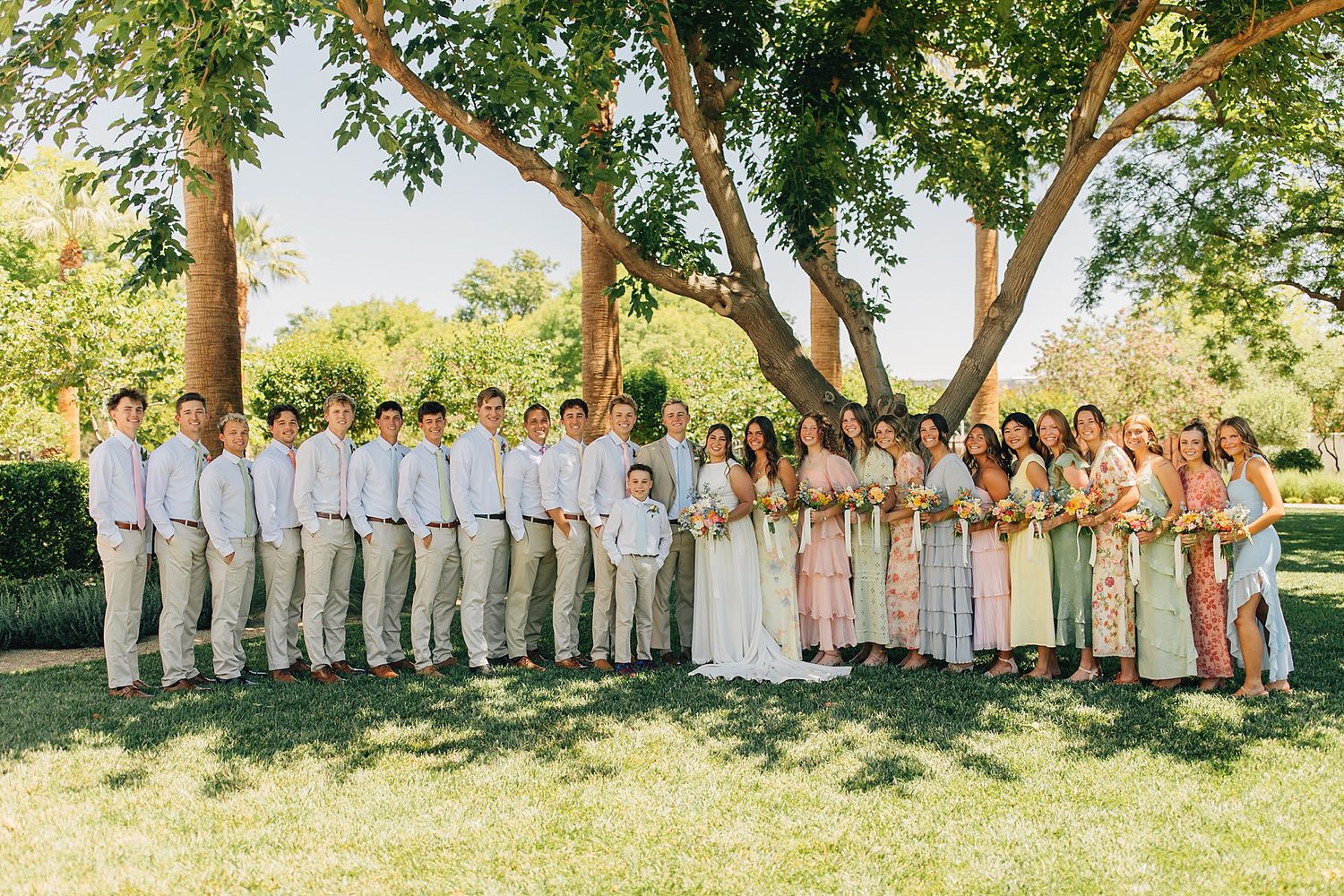 Large wedding party posing outdoors under trees in garden, groomsmen in white shirts and bridesmaids in floral dresses.