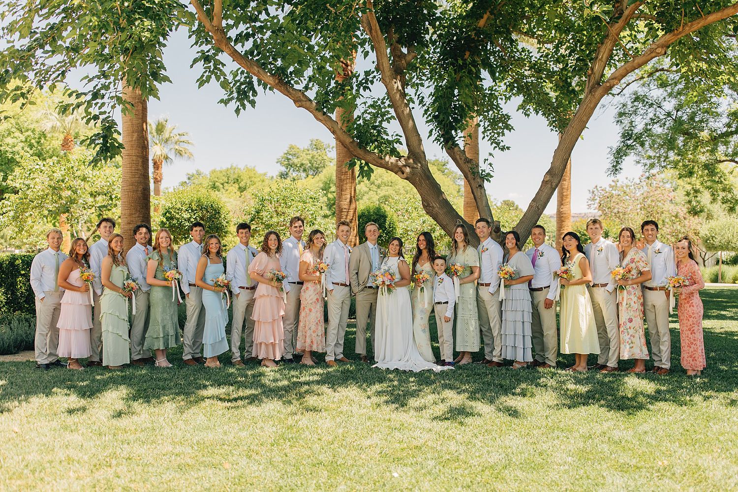Large wedding party poses outdoors under trees in coordinated pastel outfits on a sunny day.