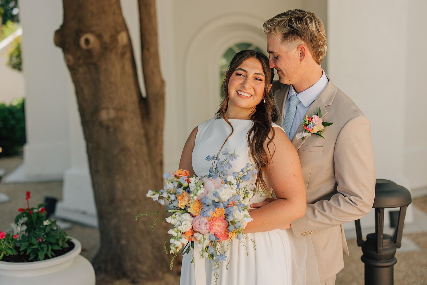 Bride and groom share a romantic moment outdoors, bride holding colorful bouquet, groom in tan suit with floral boutonniere.