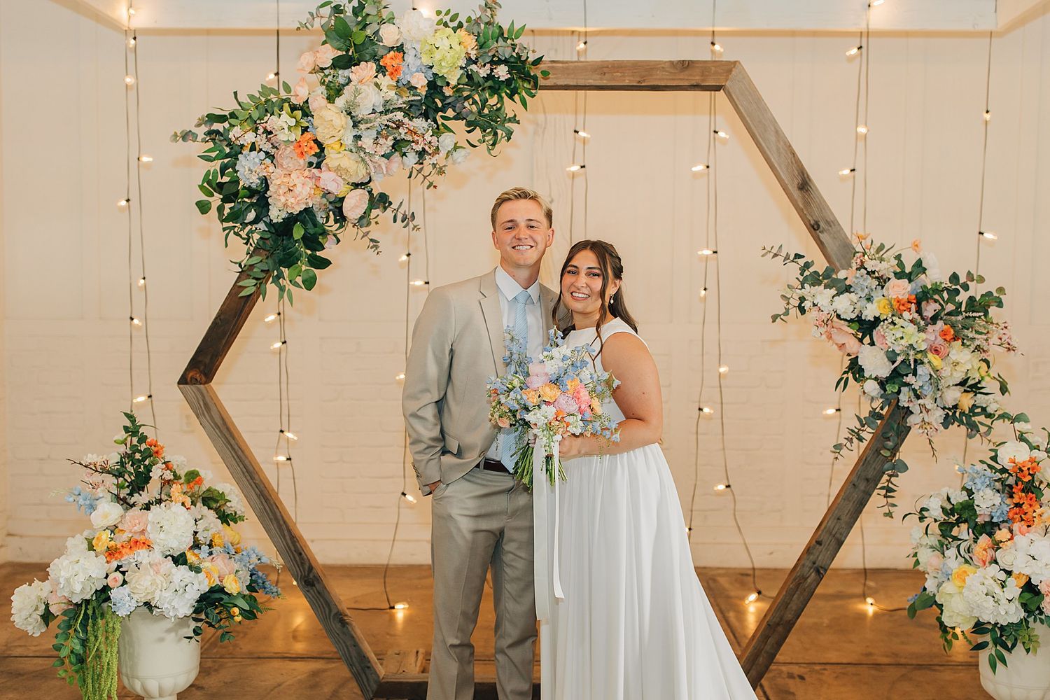 Smiling bride and groom pose under a hexagonal floral arch with string lights at an elegant wedding ceremony.