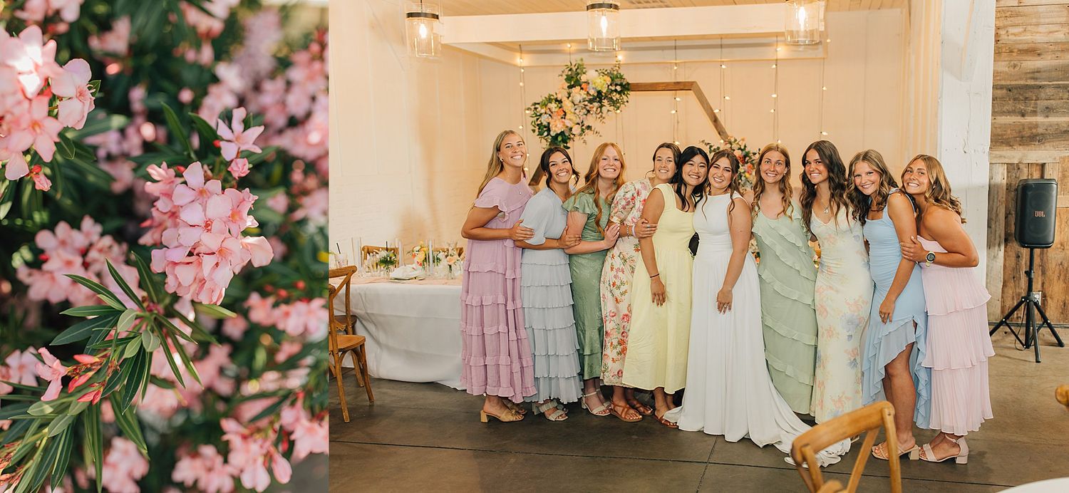 Bridesmaids in pastel dresses pose together at a wedding reception in a bright white barn venue with floral decor.