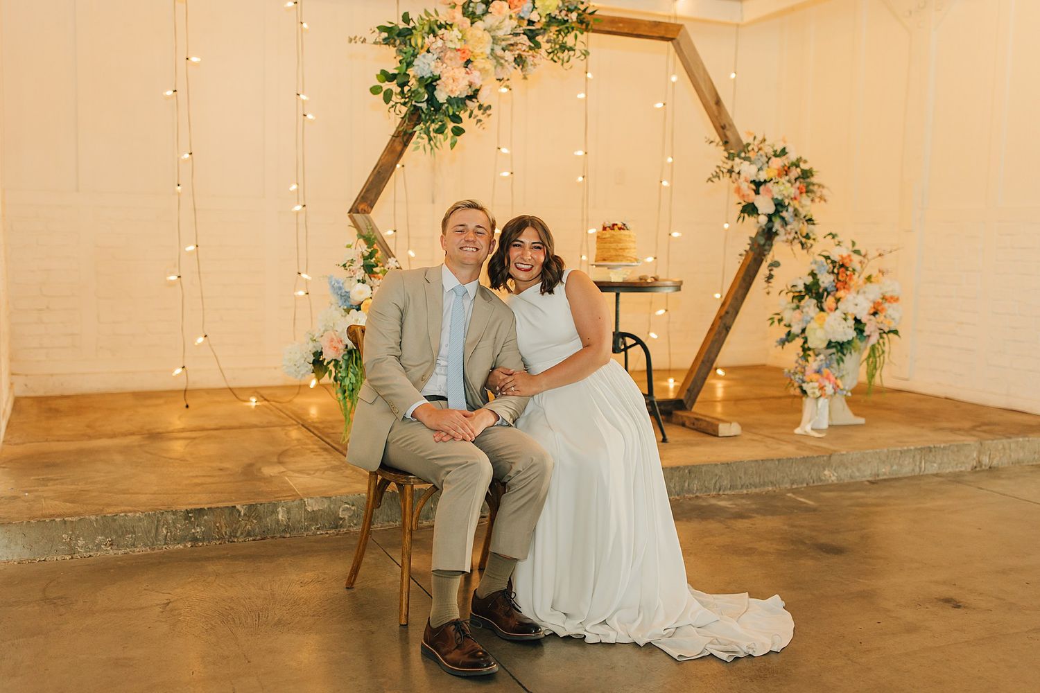 Bride and groom sitting together under a hexagonal wooden arch with floral decor and fairy lights at a wedding reception.