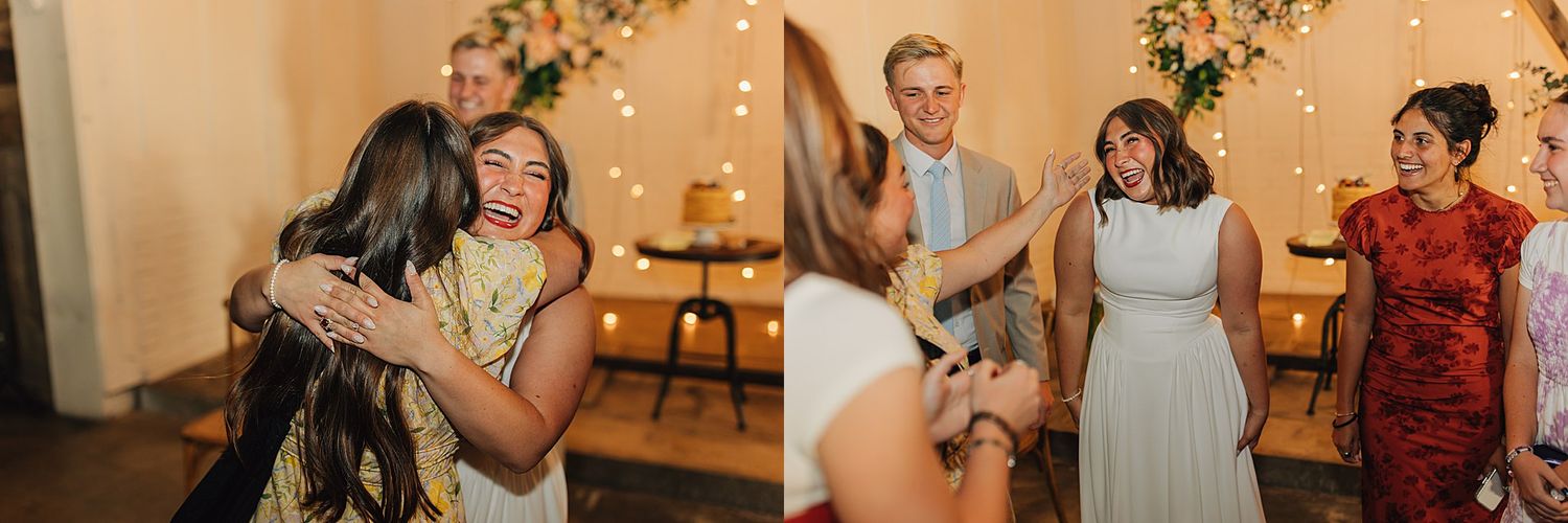 Two women share a warm hug at a wedding reception with string lights and a cake in the background.