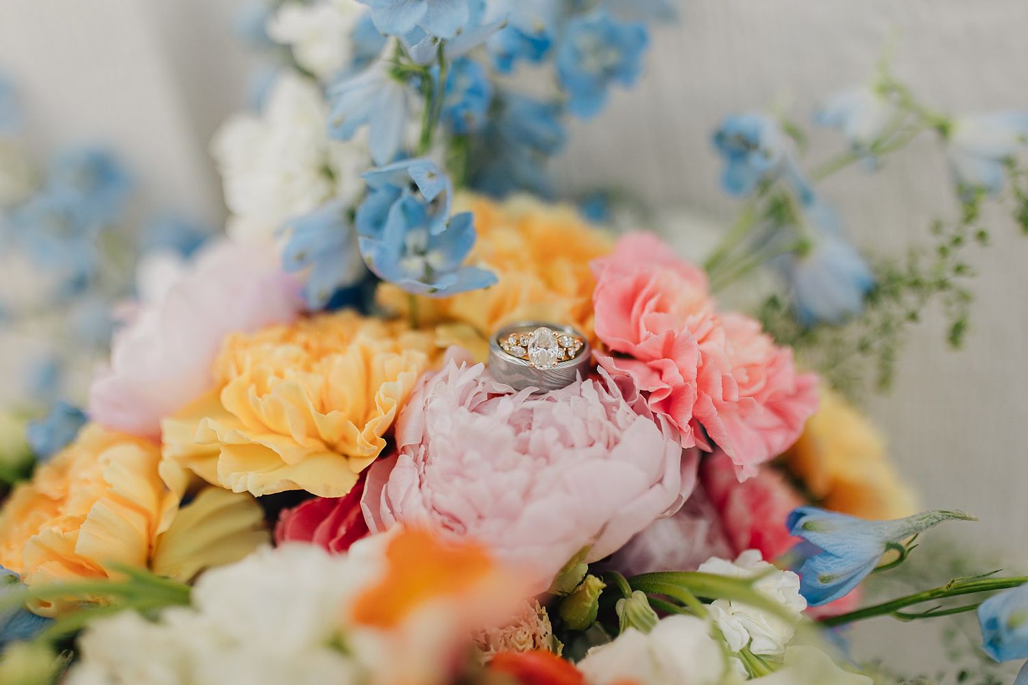 Diamond engagement ring resting on a colorful bridal bouquet with pink peonies, yellow, orange, and blue flowers.