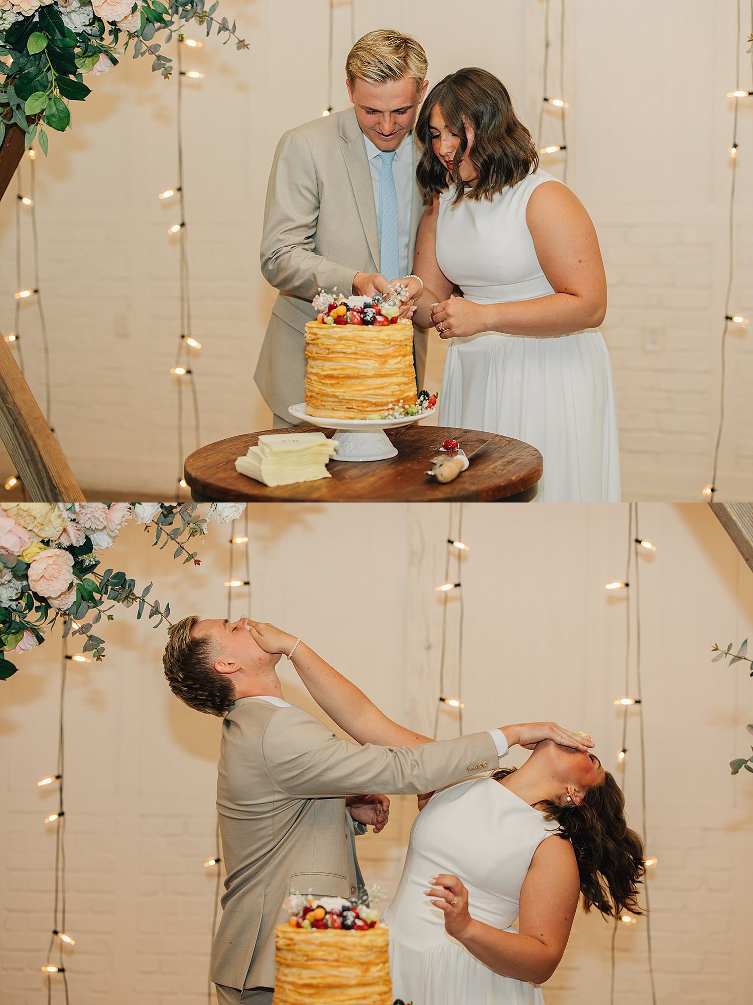 Bride and groom cutting and smashing wedding cake at rustic reception with string lights and floral backdrop.
