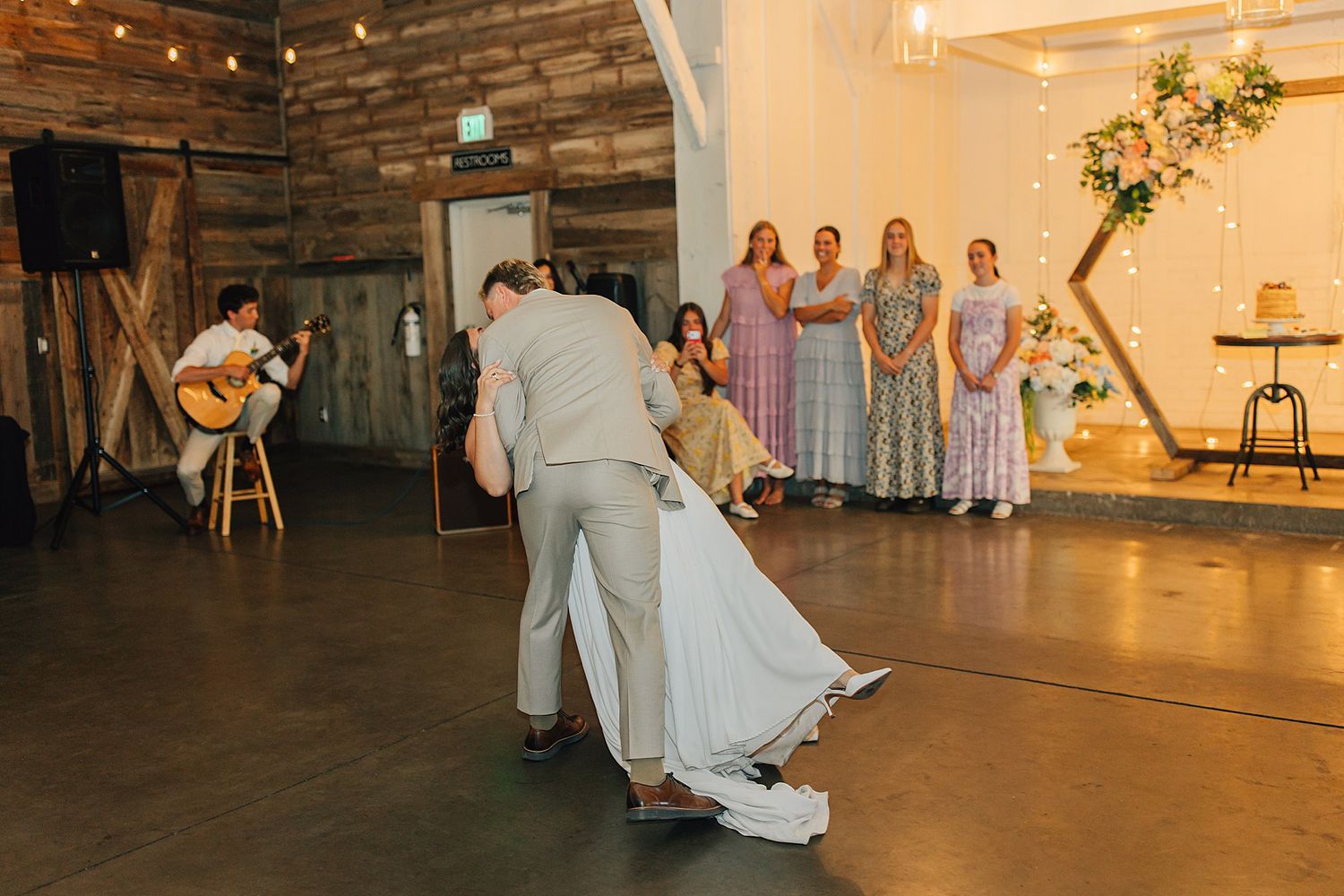 Bride and groom share first dance dip in rustic barn wedding venue with bridesmaids and live guitarist in background.