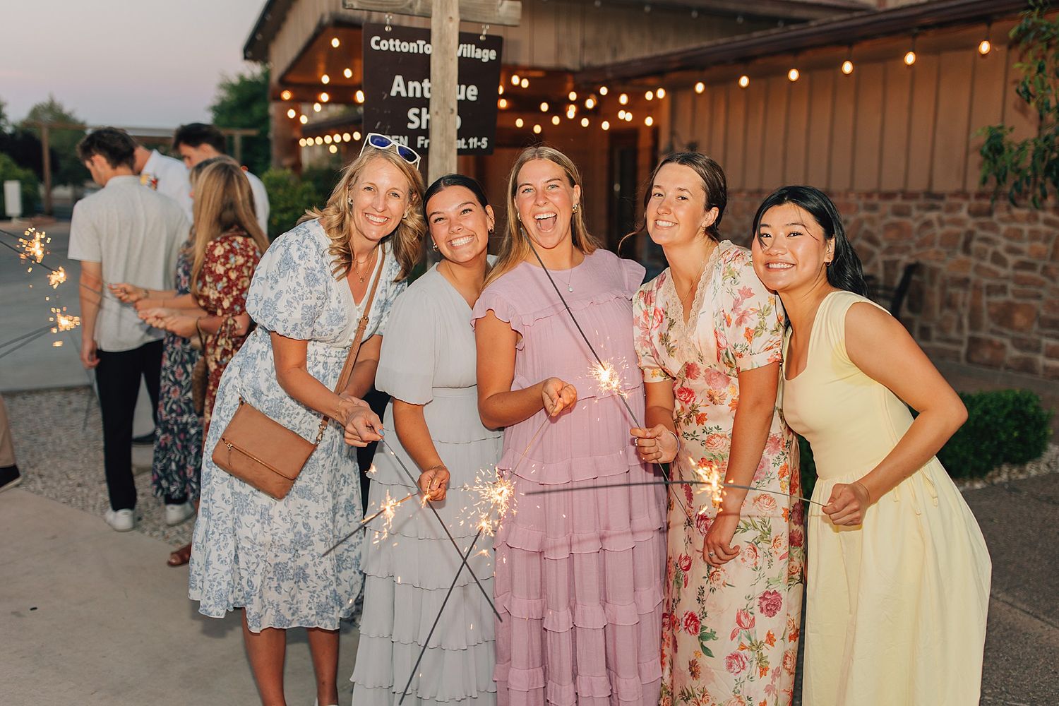 Five women smiling and holding sparklers at an outdoor evening wedding celebration near a rustic barn venue.