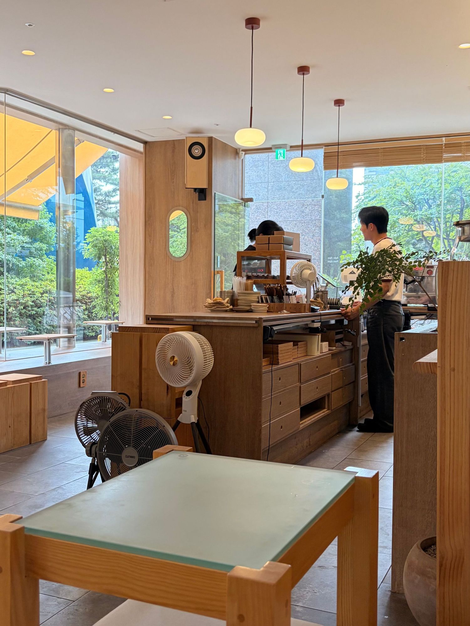 Modern Japanese cafe interior with wooden furniture, pendant lights, and large windows overlooking lush greenery.