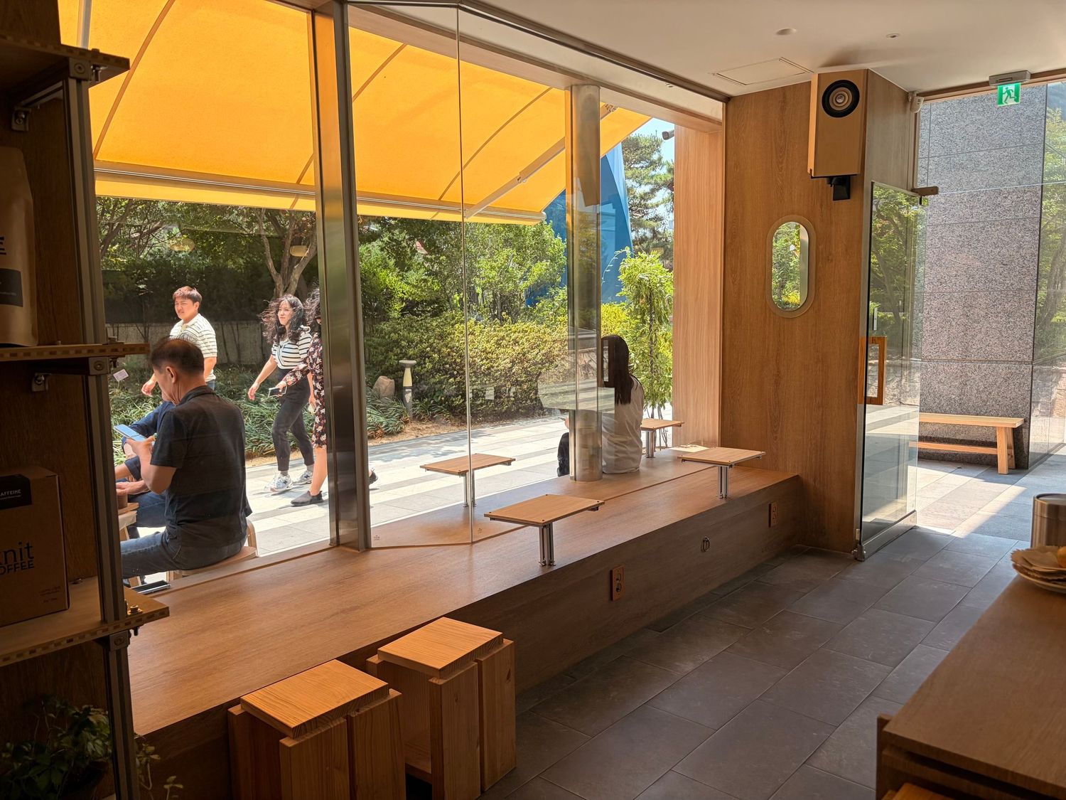 Modern cafe interior with wooden benches and tables near large windows overlooking a sunny outdoor plaza.
