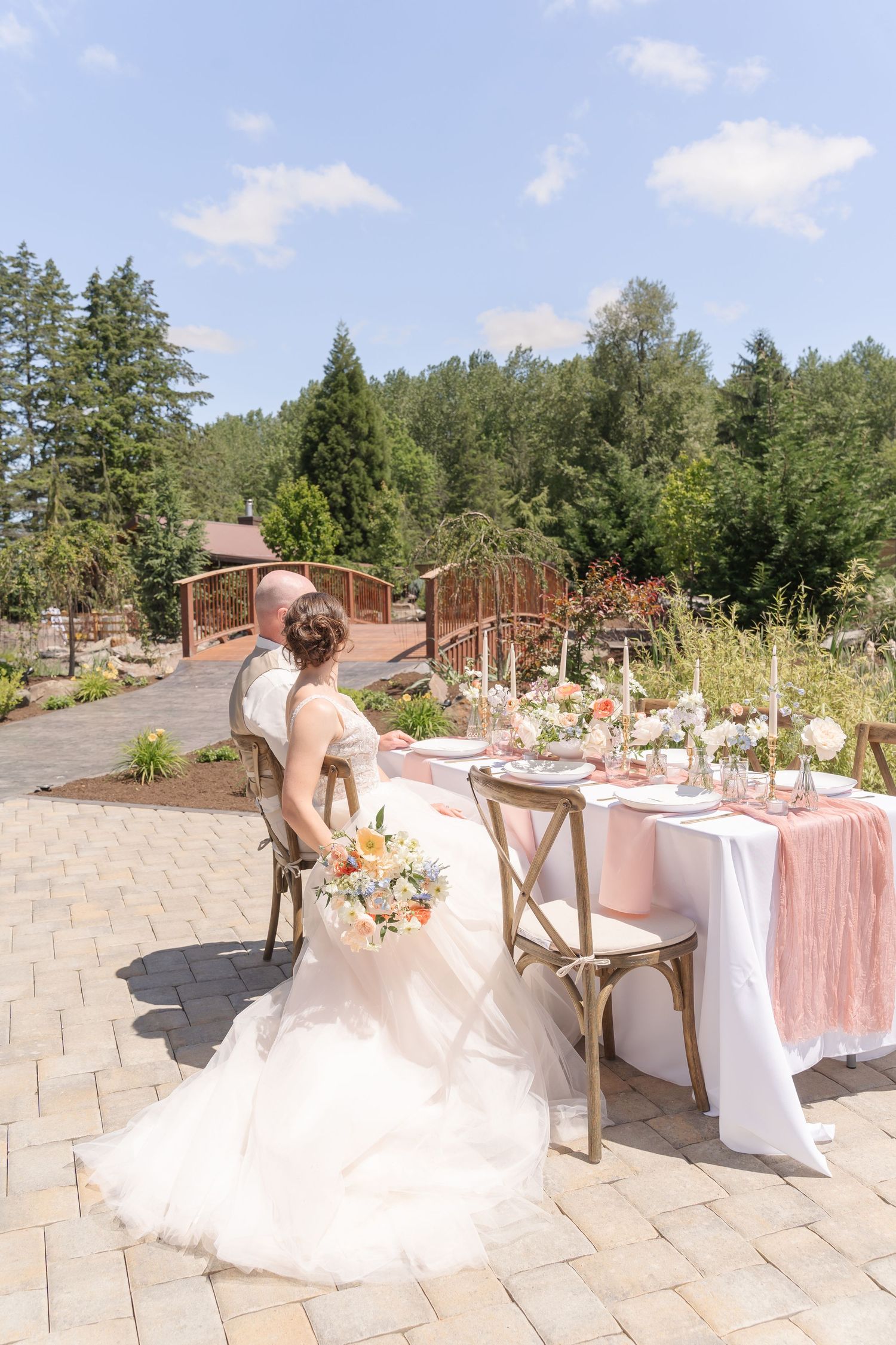 Bride and groom sit at an elegant outdoor wedding reception table with pink linens, floral arrangements, and lush forest backdrop.