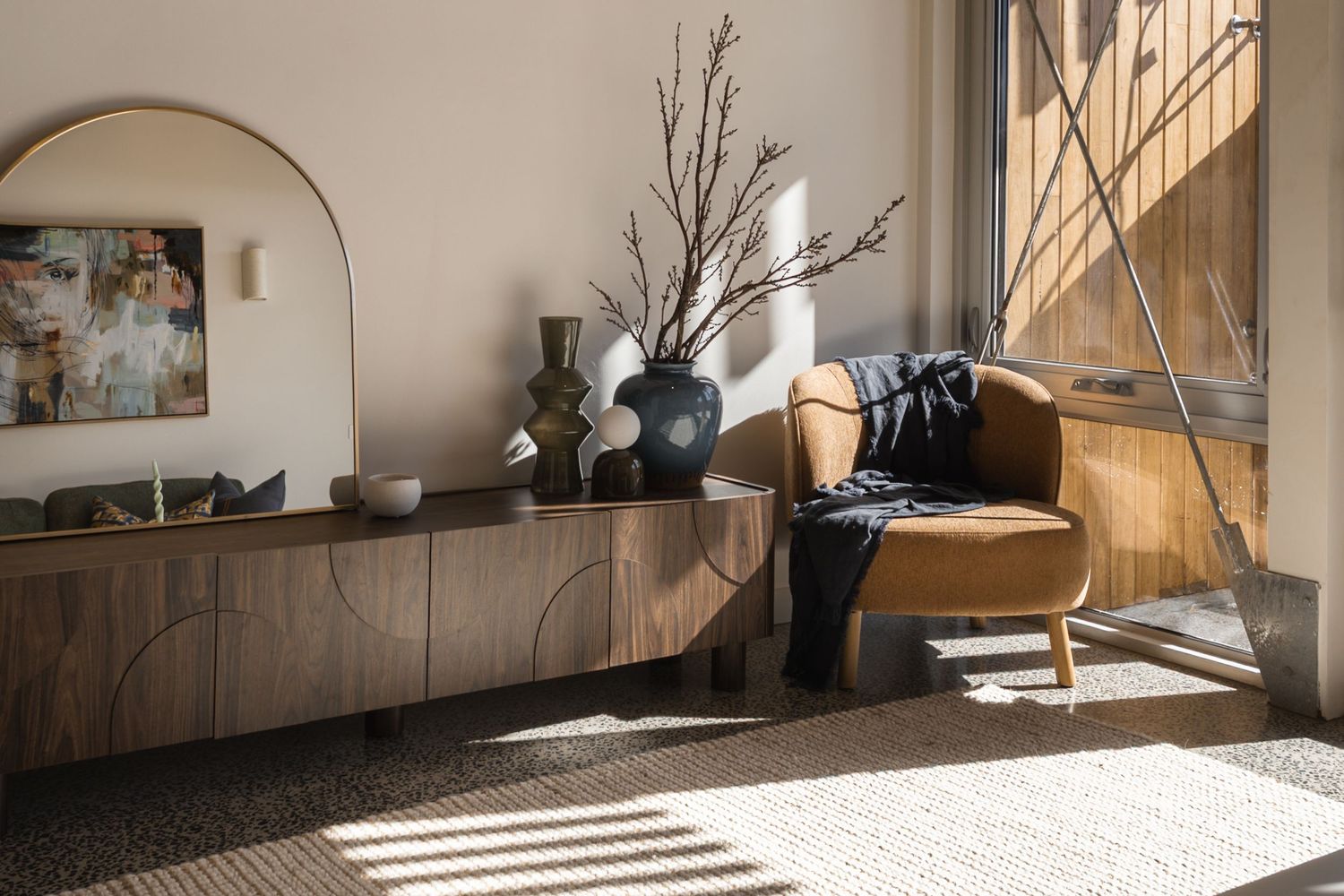 Modern living room with walnut sideboard, arched mirror, black vase with branches, and tan leather armchair by sunny window.
Middleton Field